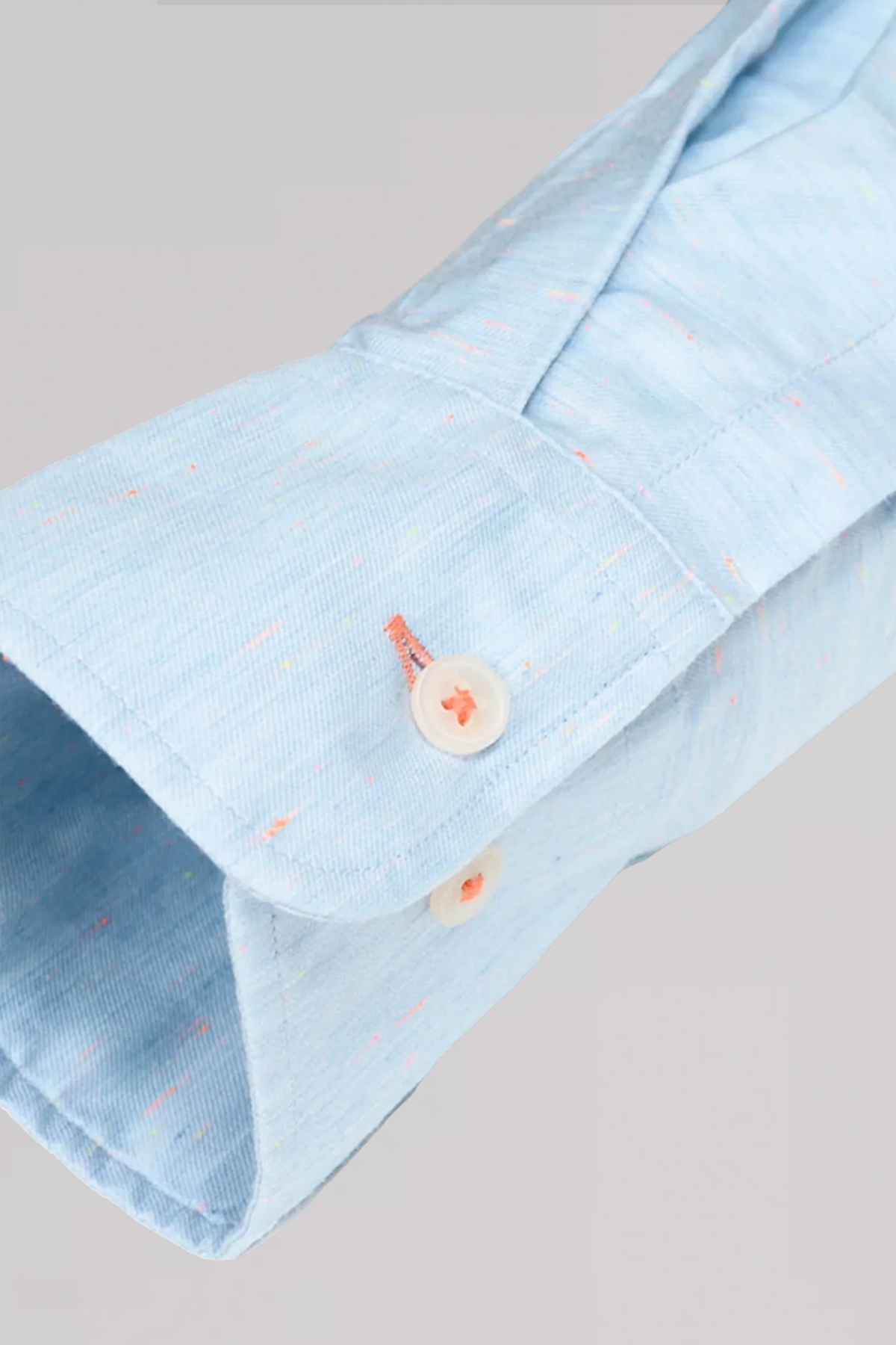 A close-up of Twillory's untuck(able) Caribbean Melange shirt sleeve in light blue with multicolored specks, rolled up to show a white button through an orange-stitched hole. Made from subtle textured, non-iron stretch fabric.