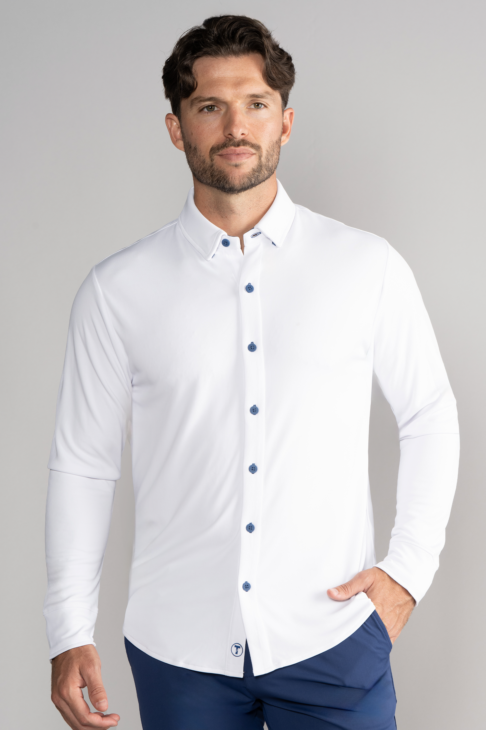 A man with short brown hair and a trimmed beard wears the Twillory Long Sleeve Button Down Polo Contrast in White Blue, paired with dark blue pants. He stands facing forward, one hand in his pocket, against a plain light gray background.