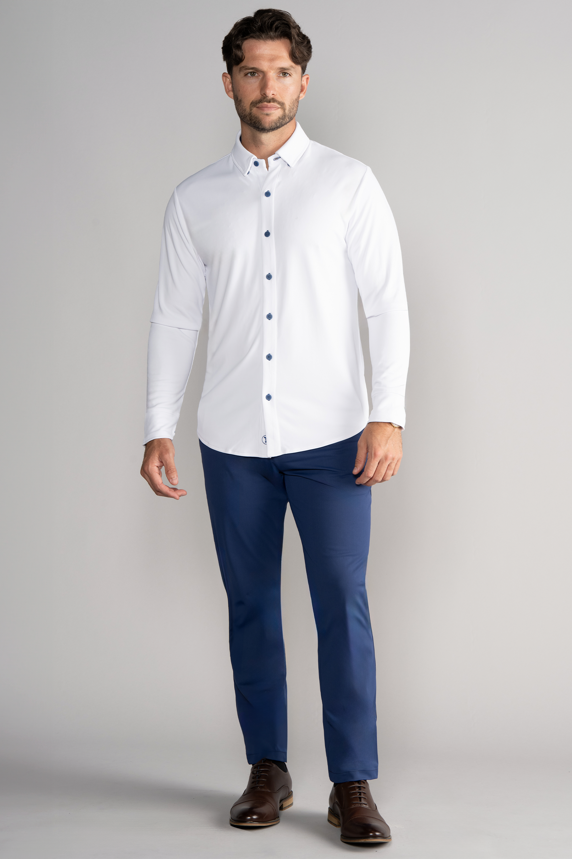A man with short brown hair and a trimmed beard stands against a light gray background, wearing the Twillory Long Sleeve Button Down Polo Contrast in White Blue, blue dress pants, and brown leather dress shoes, facing the camera neutrally.