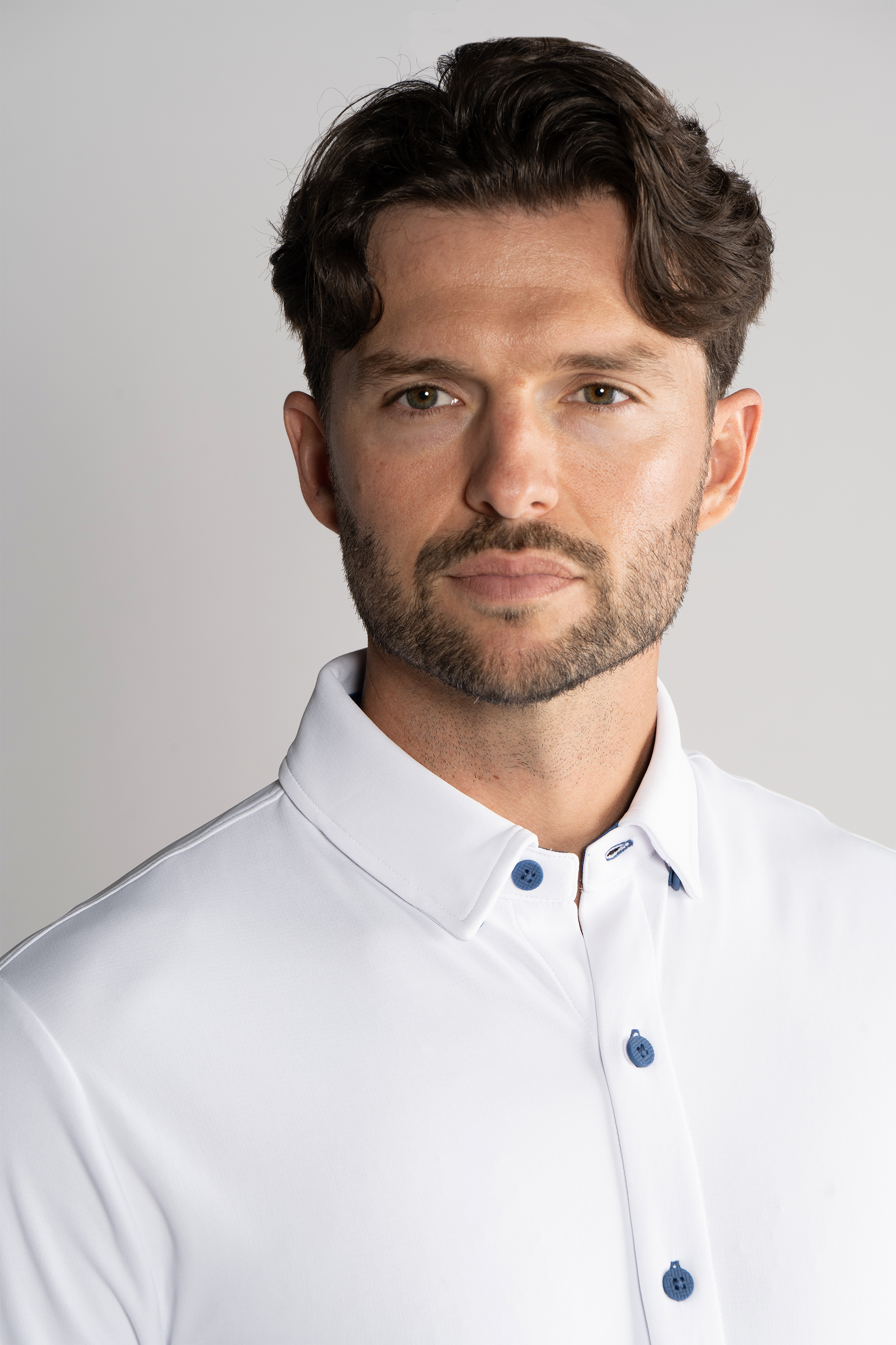 A man with wavy dark hair and a trimmed beard wears the Twillory Long Sleeve Button Down Polo Contrast in white/blue. He faces the camera against a plain light gray background, with only his upper torso and head visible, and a neutral expression.