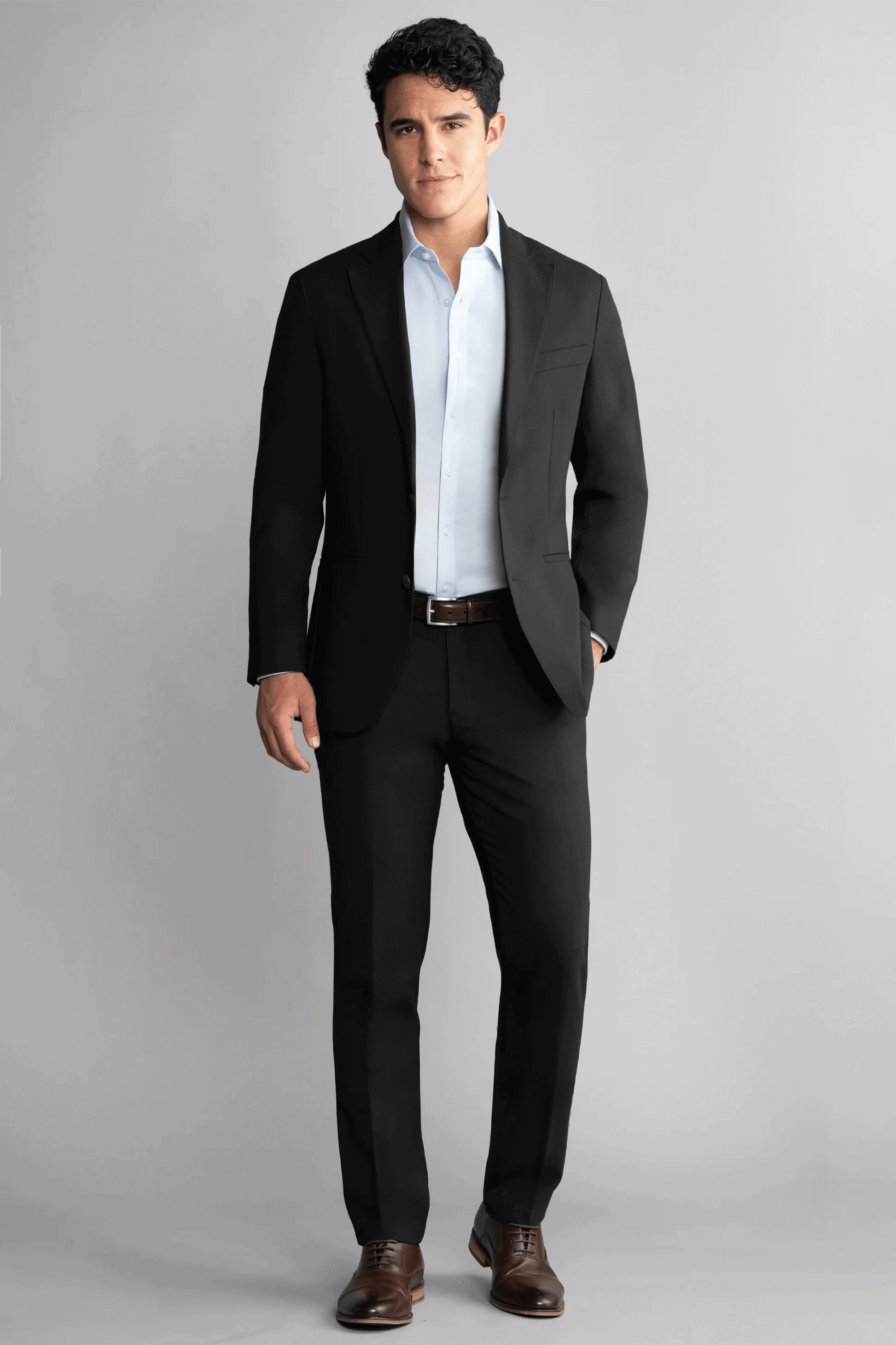 A man stands against a plain light gray background wearing the Twillory Hybrid Wool Blazer in black over a light blue shirt and brown dress shoes, left hand in pocket, confidently facing the camera with a neutral expression.