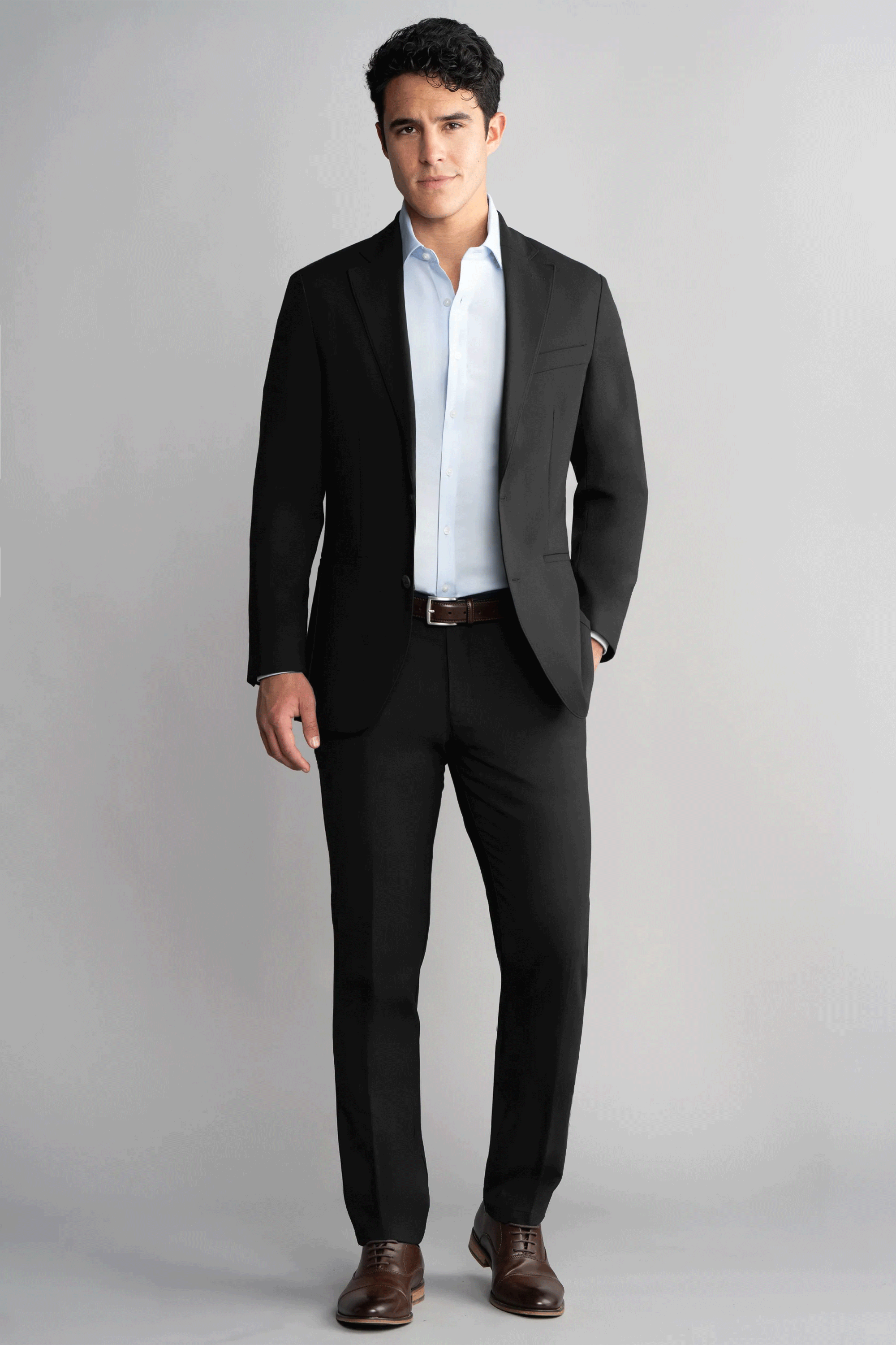 A man with short dark hair stands against a plain gray background, facing forward with a neutral expression. He wears Twillory’s Hybrid Wool Pants in black, paired with a light blue dress shirt and brown shoes, one hand in his pocket.