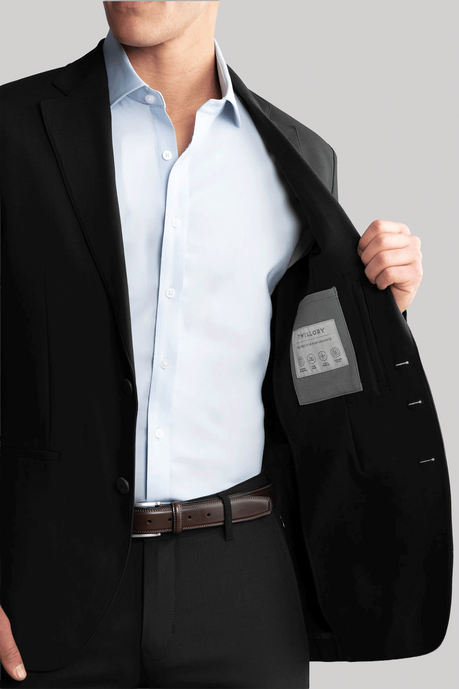 A man in a Twillory Hybrid Wool Blazer // Black and light blue dress shirt opens his jacket to show the inner pocket and label. He wears a brown leather belt, with his face partly out of frame against a plain light gray background.
