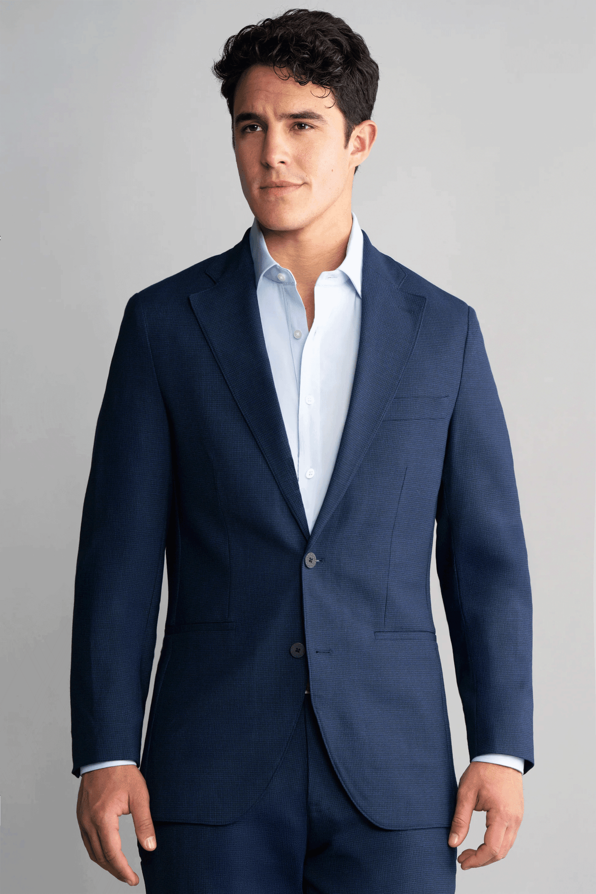 A man with short curly dark hair models the Twillory Hybrid Wool Blazer in Navy Puppytooth over a light blue shirt, standing against a plain light gray backdrop and looking slightly to the side with a neutral expression.