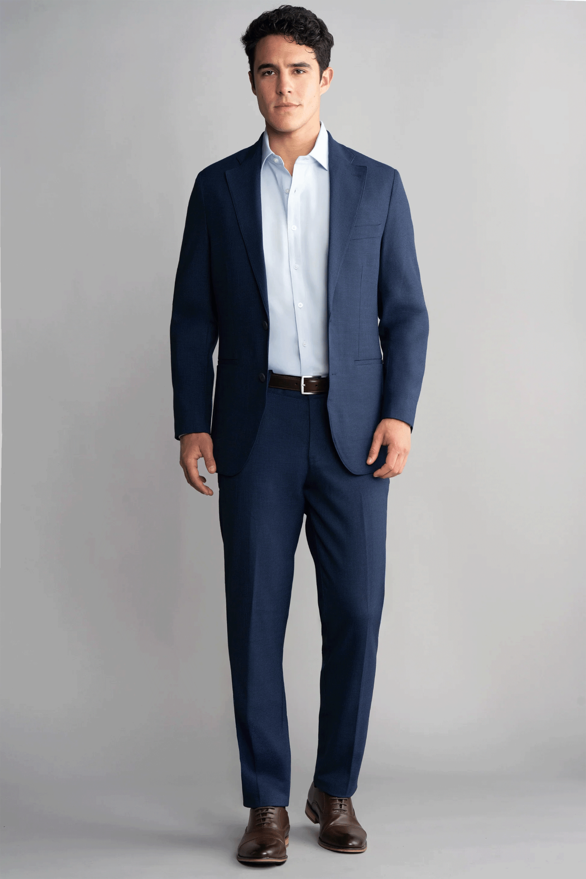 A man stands facing forward against a light gray background, wearing Twillory Hybrid Wool Pants in Navy Puppytooth with a light blue shirt, dark belt, and brown dress shoes. His short dark hair and relaxed posture finish his modern look.