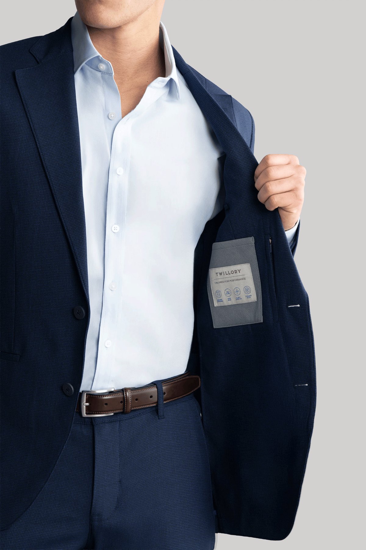 A man in a Navy Puppytooth Hybrid Wool Blazer by Twillory stands against a gray background, holding open his jacket to reveal the branded inner pocket. His stylish blazer features 4-way stretch and is machine washable. His head is partially out of frame.