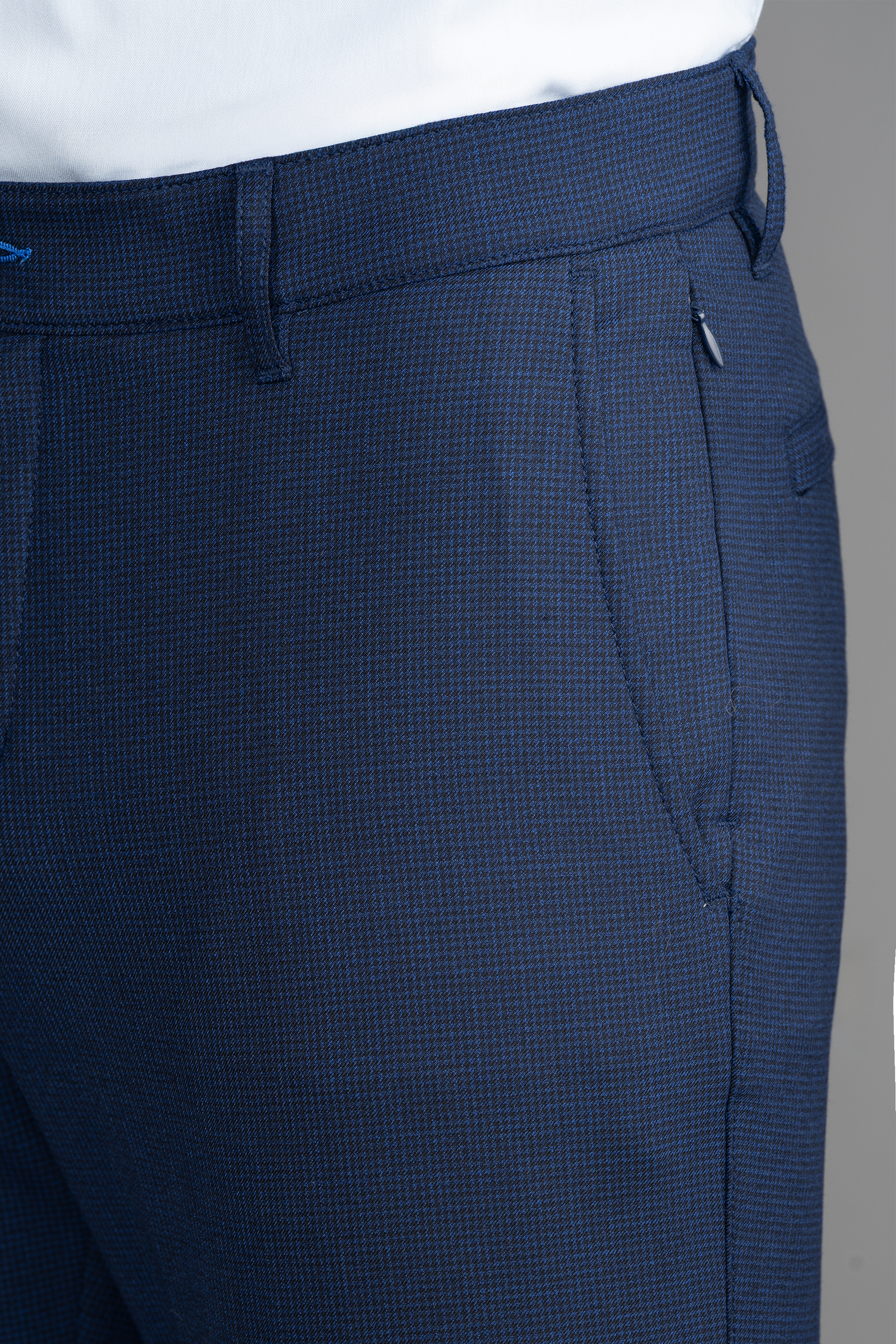 Close-up of Twillory Hybrid Wool Pants // Navy Puppytooth, featuring a subtle check pattern and 4-way stretch. A white shirt is tucked in, with the person’s right hand in the pocket. Plain gray background.