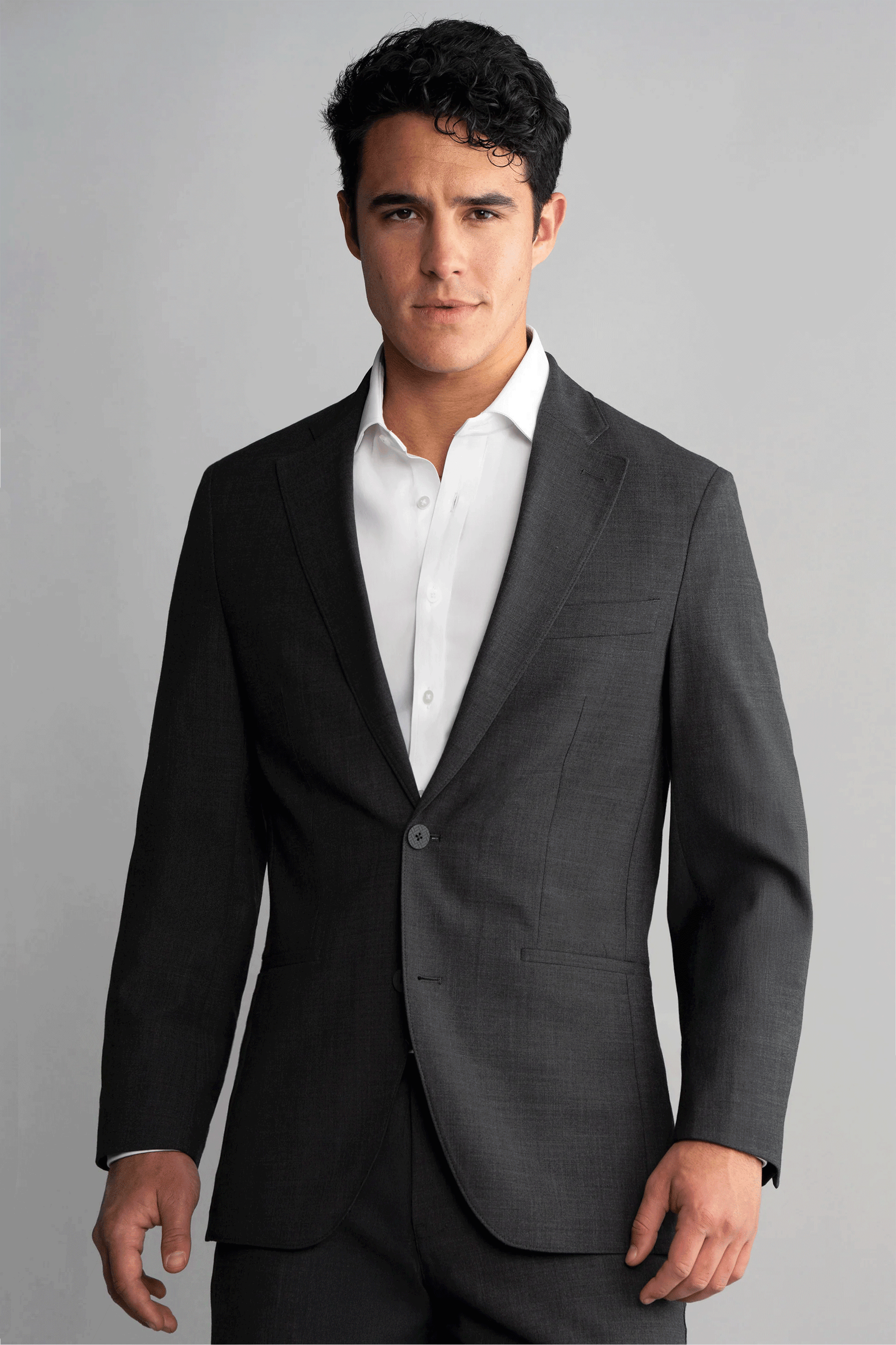 A man with short, dark, wavy hair wears the Twillory Hybrid Wool Blazer in Grey Heather with matching pants and a white shirt. He stands against a light gray background, looking straight ahead with a neutral expression.