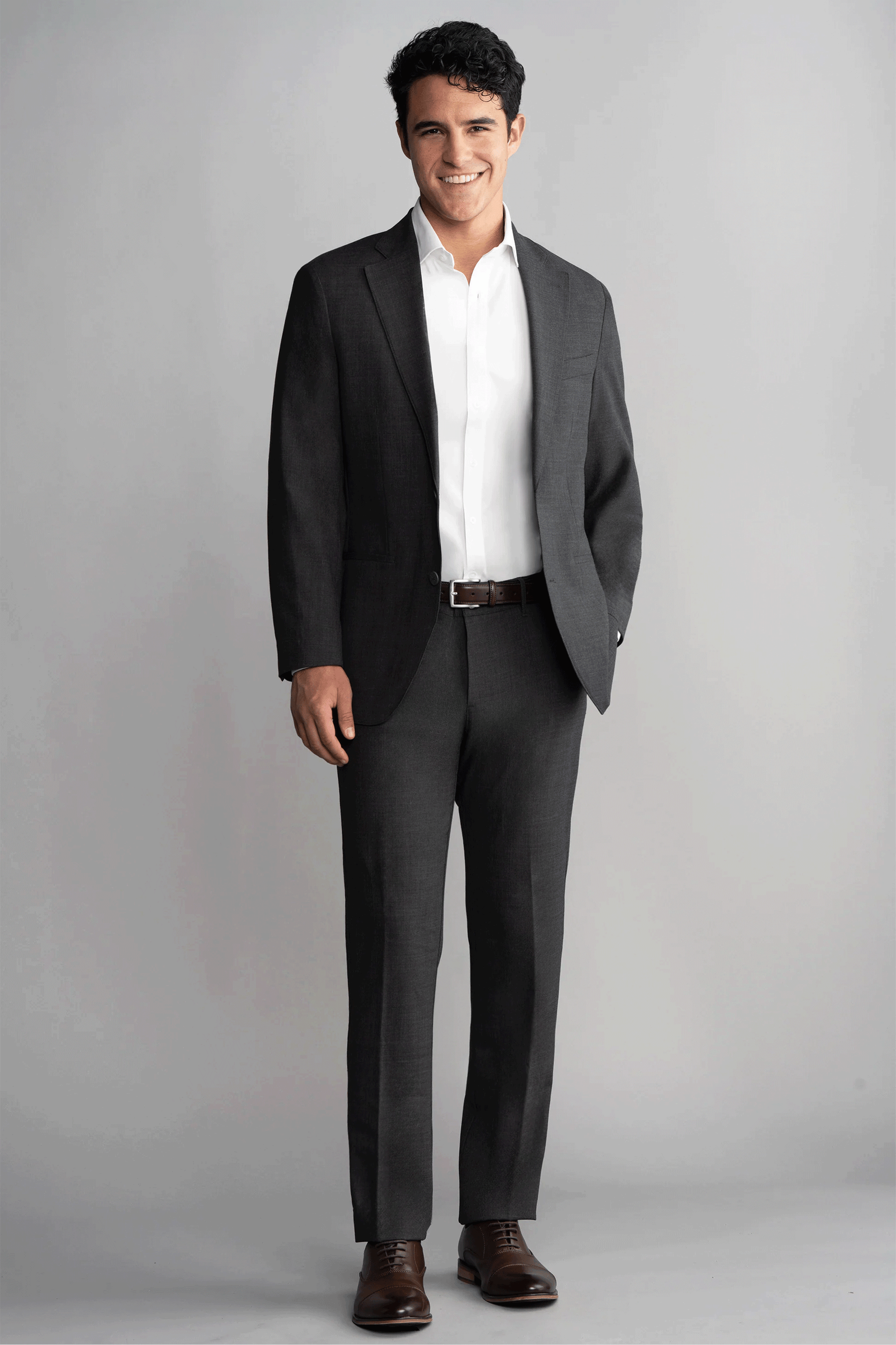 A man smiles against a light gray background, wearing Twillory’s Hybrid Wool Blazer in Grey Heather with a white shirt, brown belt, and dress shoes. His left hand is in his pocket and his dark hair is neatly styled.