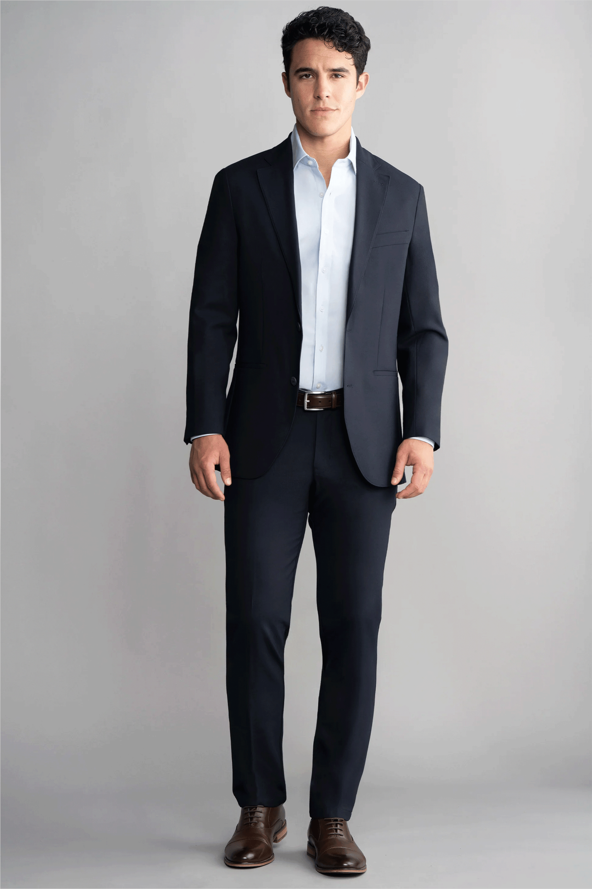 A man stands facing forward before a light gray background. He wears the Twillory Hybrid Wool Blazer in Navy, paired with a white dress shirt, brown belt, and brown shoes. His expression is neutral and his hands rest at his sides.