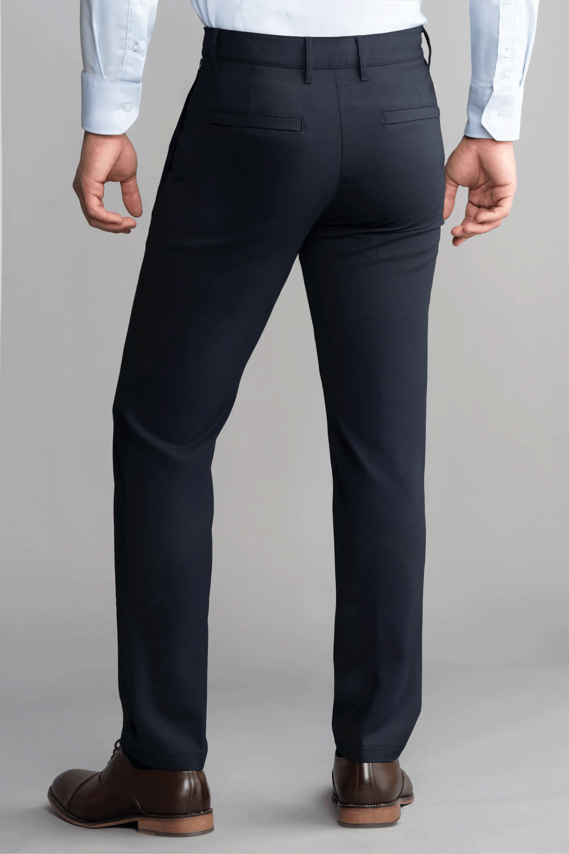 A person wears Twillory Hybrid Wool Pants in navy and a light blue button-down shirt with brown dress shoes, standing relaxed against a light gray background. The pants feature two back pockets with buttoned flaps.