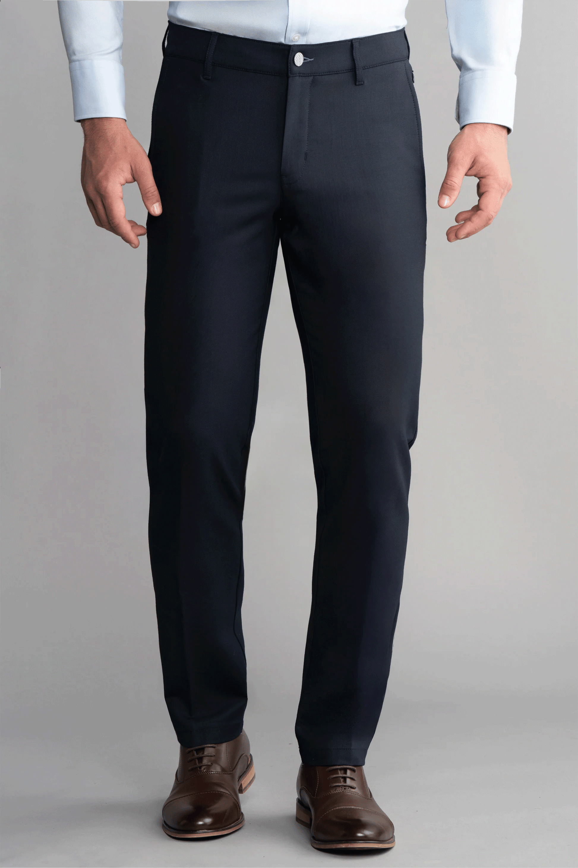 A man stands facing forward wearing Twillory Hybrid Wool Pants in Navy, a light blue dress shirt tucked in, and brown leather dress shoes. The background is plain gray. His hands are relaxed by his sides and his face is not visible.