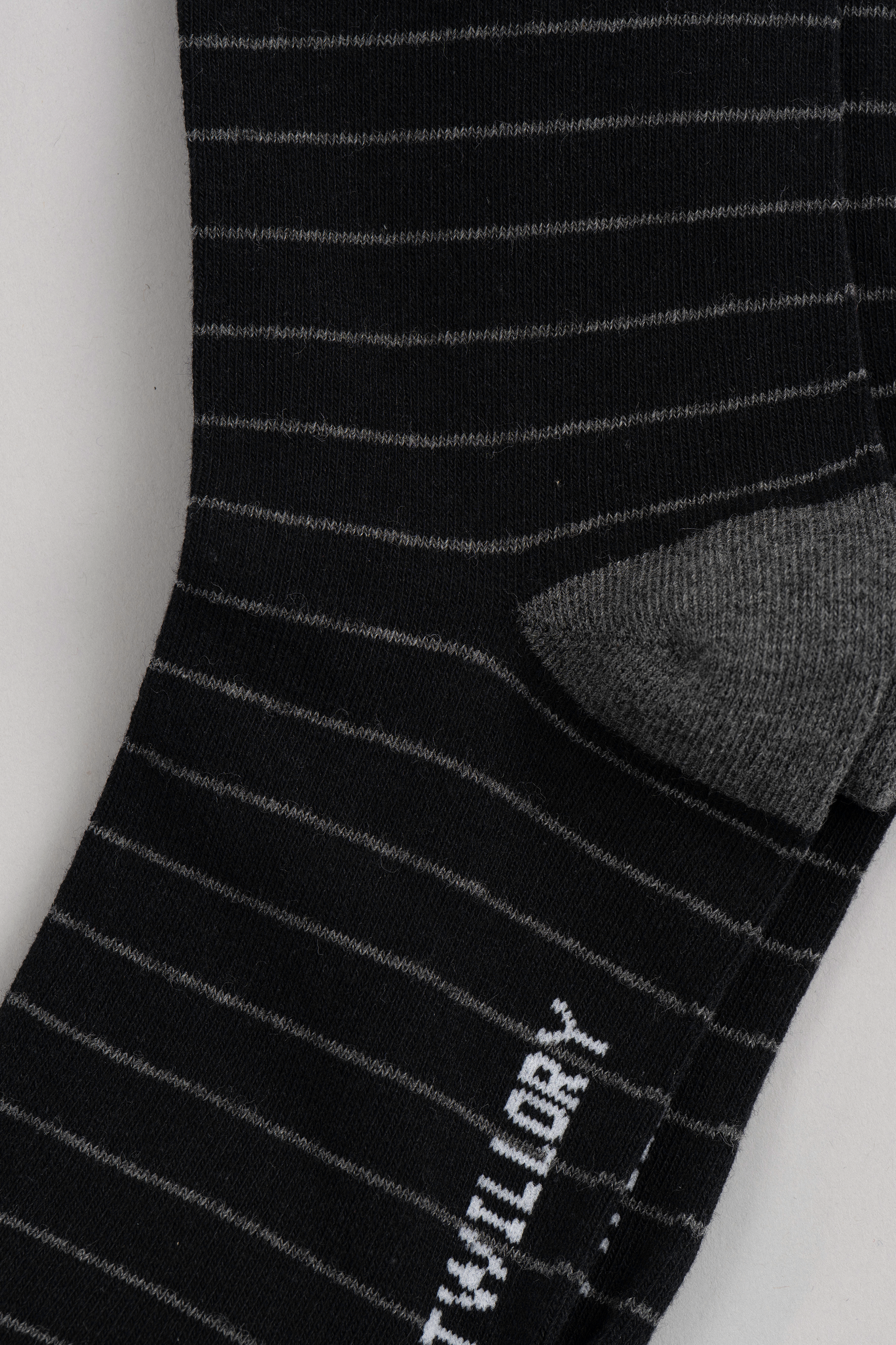 A close-up of Twillory’s Black Stripe sock, showing thin white stripes on black fabric with a solid gray heel. White text appears partially on the striped sole. The flat-laid sock rests on a light gray background.
