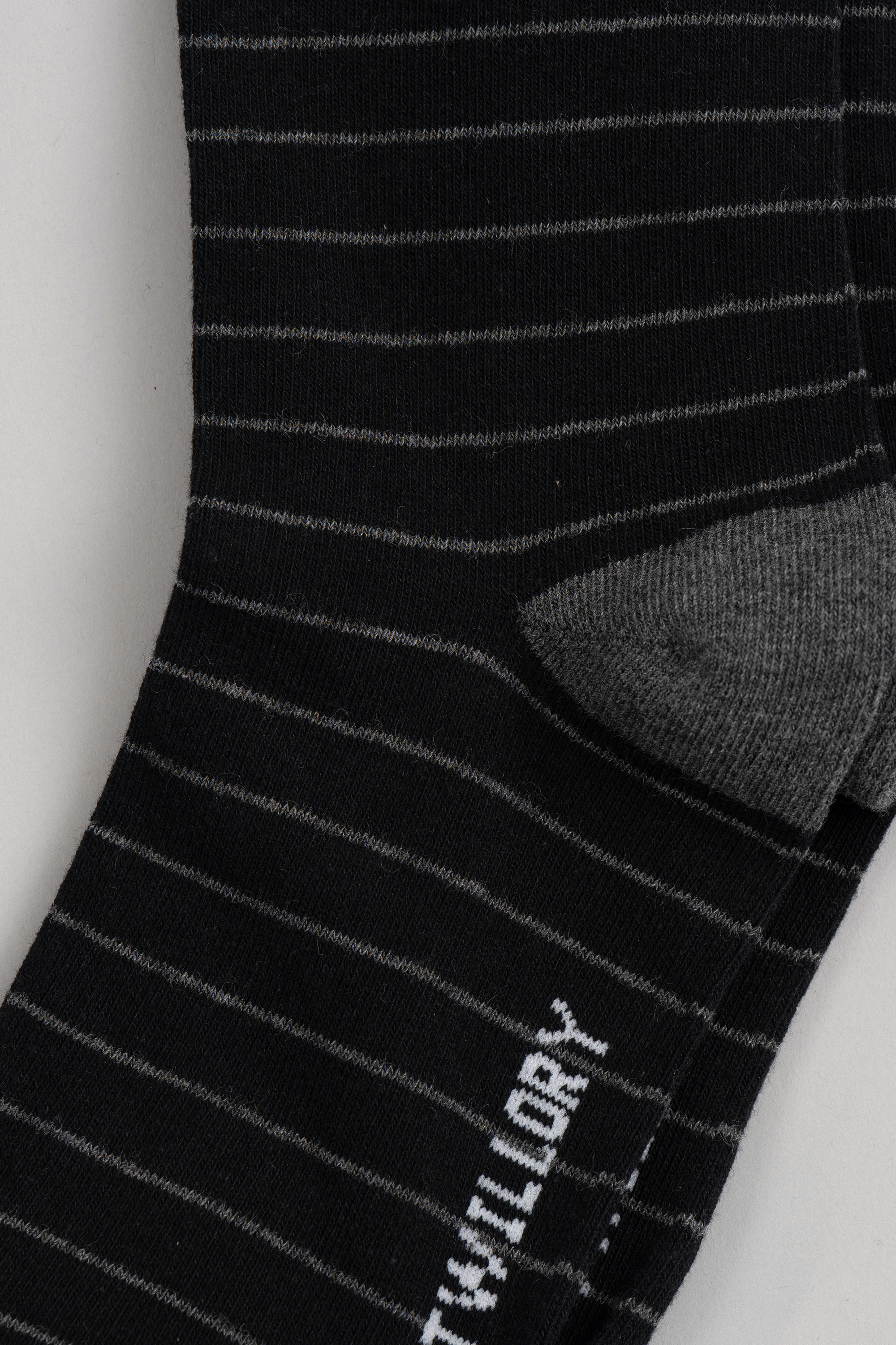 A close-up of Twillory’s Black Stripe sock, showing thin white stripes on black fabric with a solid gray heel. White text appears partially on the striped sole. The flat-laid sock rests on a light gray background.