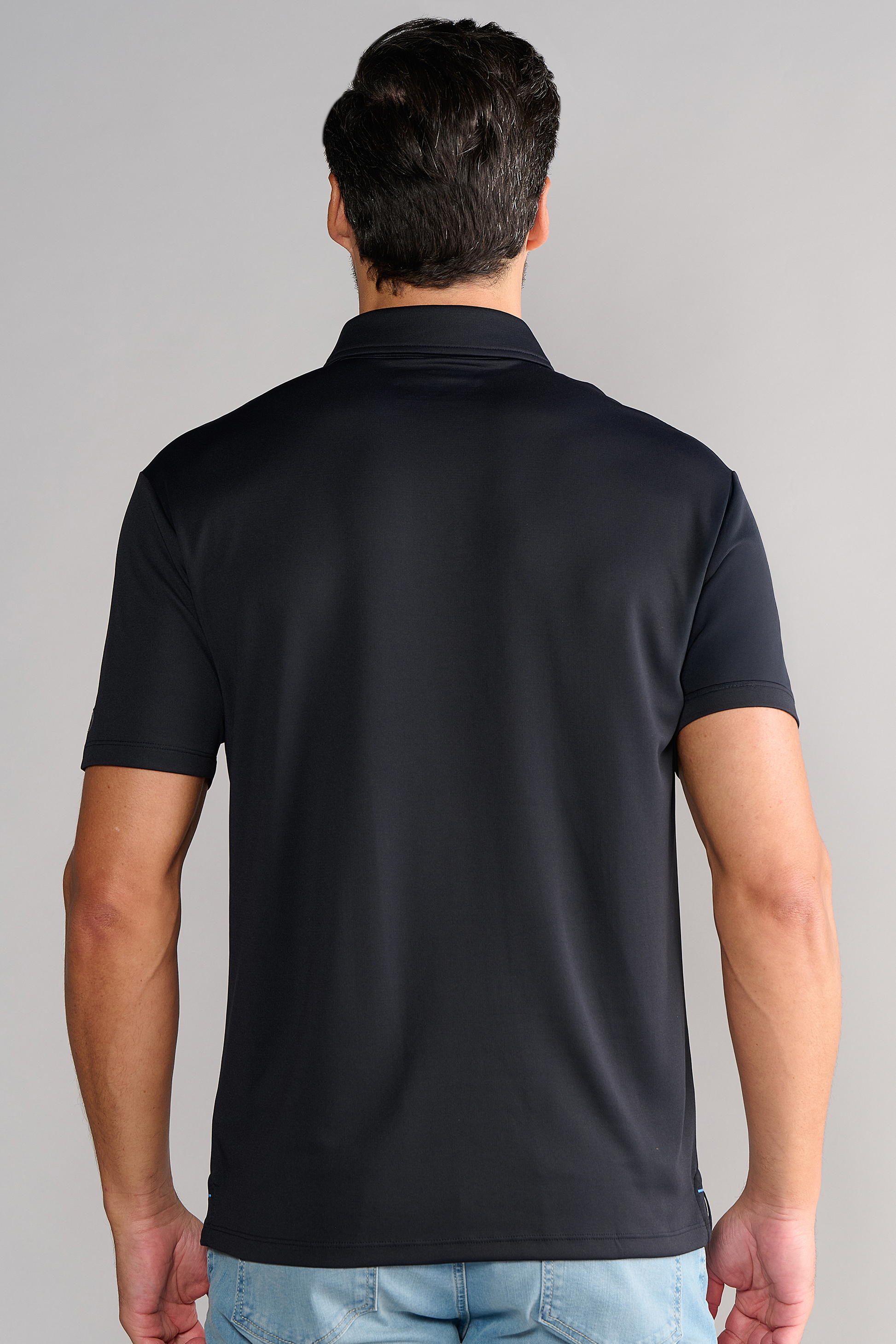 A man with short dark hair is seen from behind, standing upright in front of a plain gray background, wearing Twillory's Short Sleeve Polo Solid in black paired with light blue jeans.