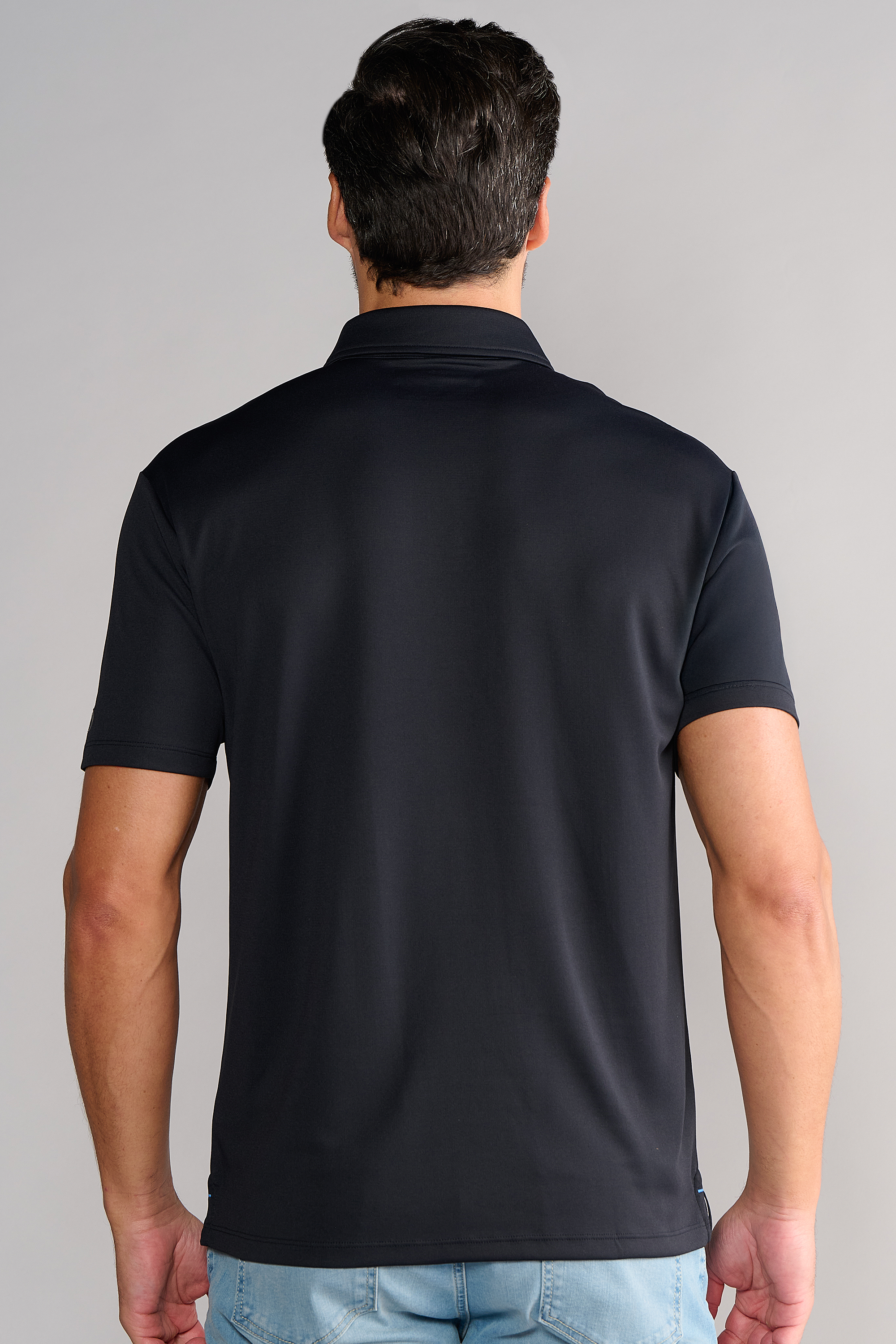 A man with short dark hair is seen from behind, standing upright in front of a plain gray background, wearing Twillory's Short Sleeve Polo Solid in black paired with light blue jeans.