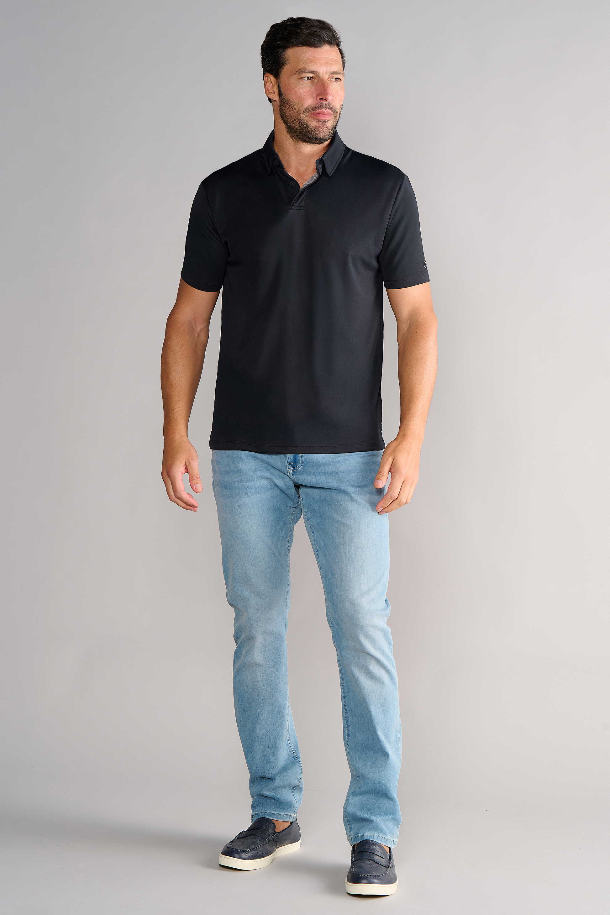 A man with short dark hair and a trimmed beard poses against a light gray background, wearing the Twillory Short Sleeve Polo Solid in black, paired with light blue jeans and dark slip-on shoes. He faces slightly left with a neutral expression.