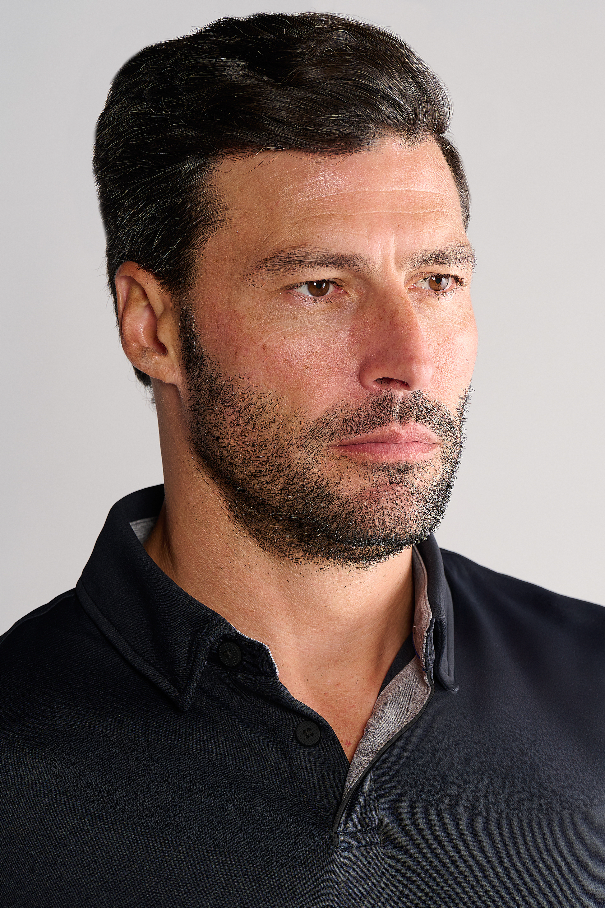 A man with short dark hair and a trimmed beard wears the Twillory Short Sleeve Polo Solid in black. The tailored-fit shirt features a grey inner lining. He looks slightly to the side with a neutral expression against a plain, light background.