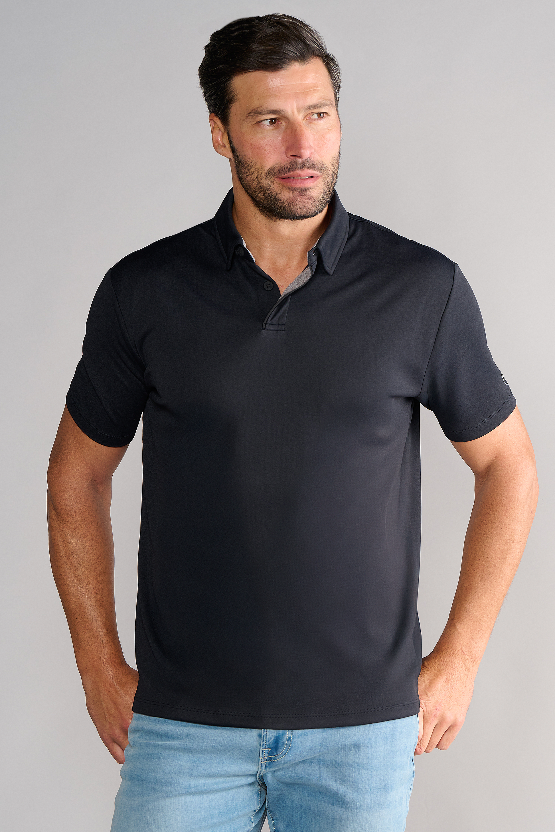 A man with short dark hair and a trimmed beard stands against a plain background, wearing Twillory's Short Sleeve Polo Solid in black and light blue jeans. His hands rest near his hips as he looks slightly to the side with a neutral expression.