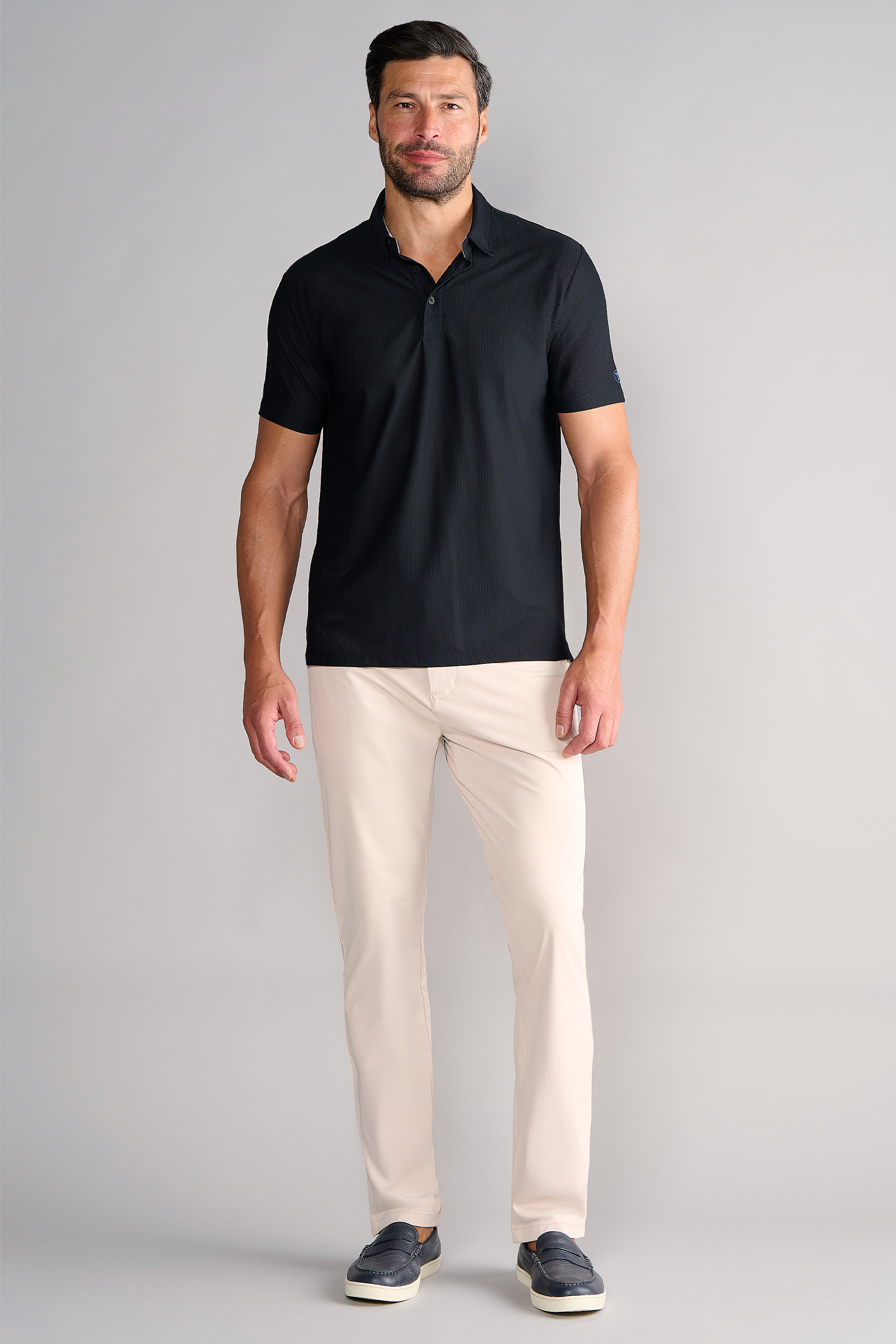 A man with short dark hair and a beard stands against a light gray background, wearing Twillory's AIR 3 Button Polo Solid in Black, paired with light beige pants and dark slip-on shoes. He smiles slightly with his arms relaxed by his sides.