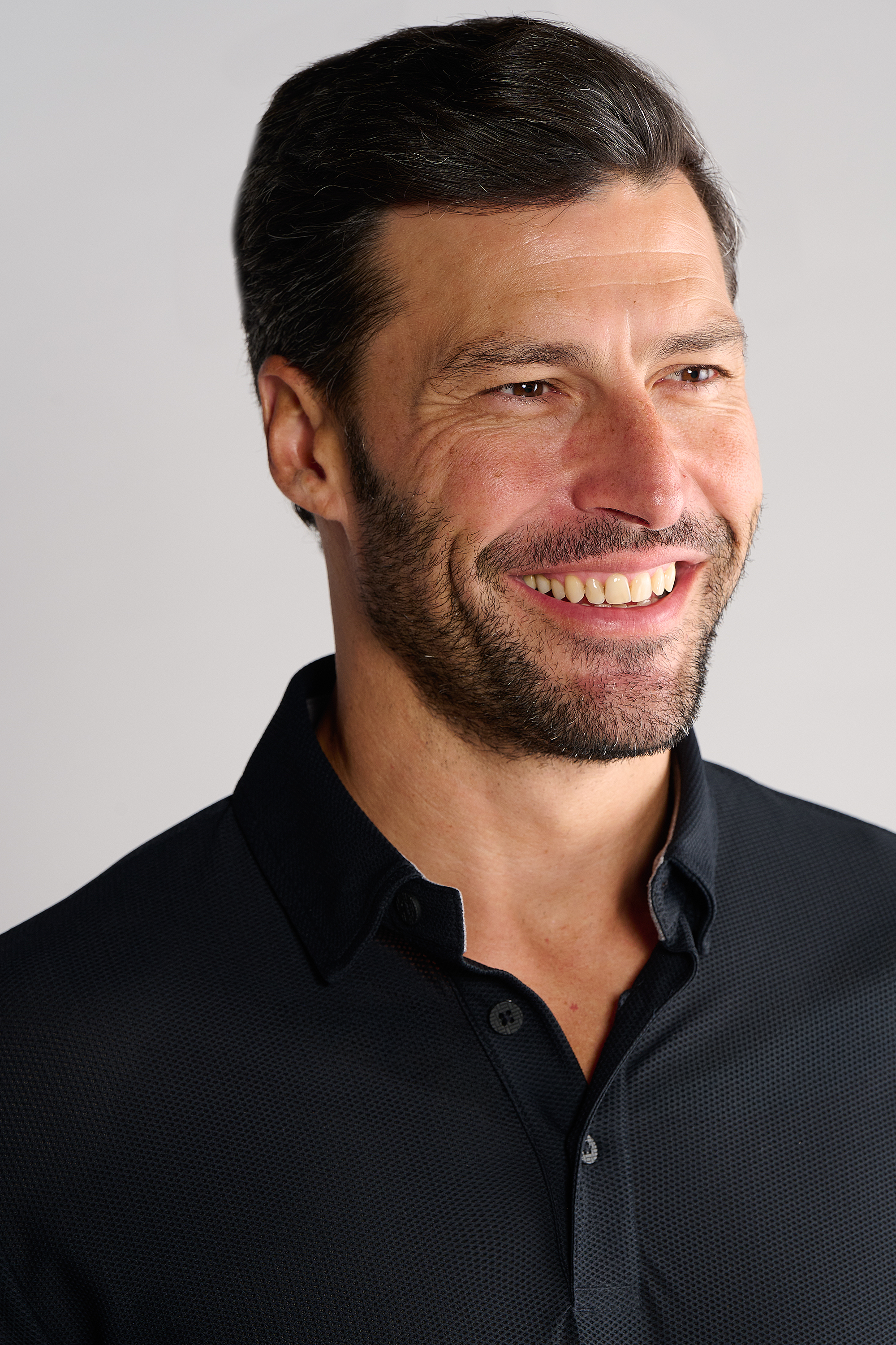 A man with short dark hair and a trimmed beard smiles, looking slightly to the right. He wears the Twillory AIR 3 Button Polo Solid in black, set against a light background. Only his upper torso and head are visible.