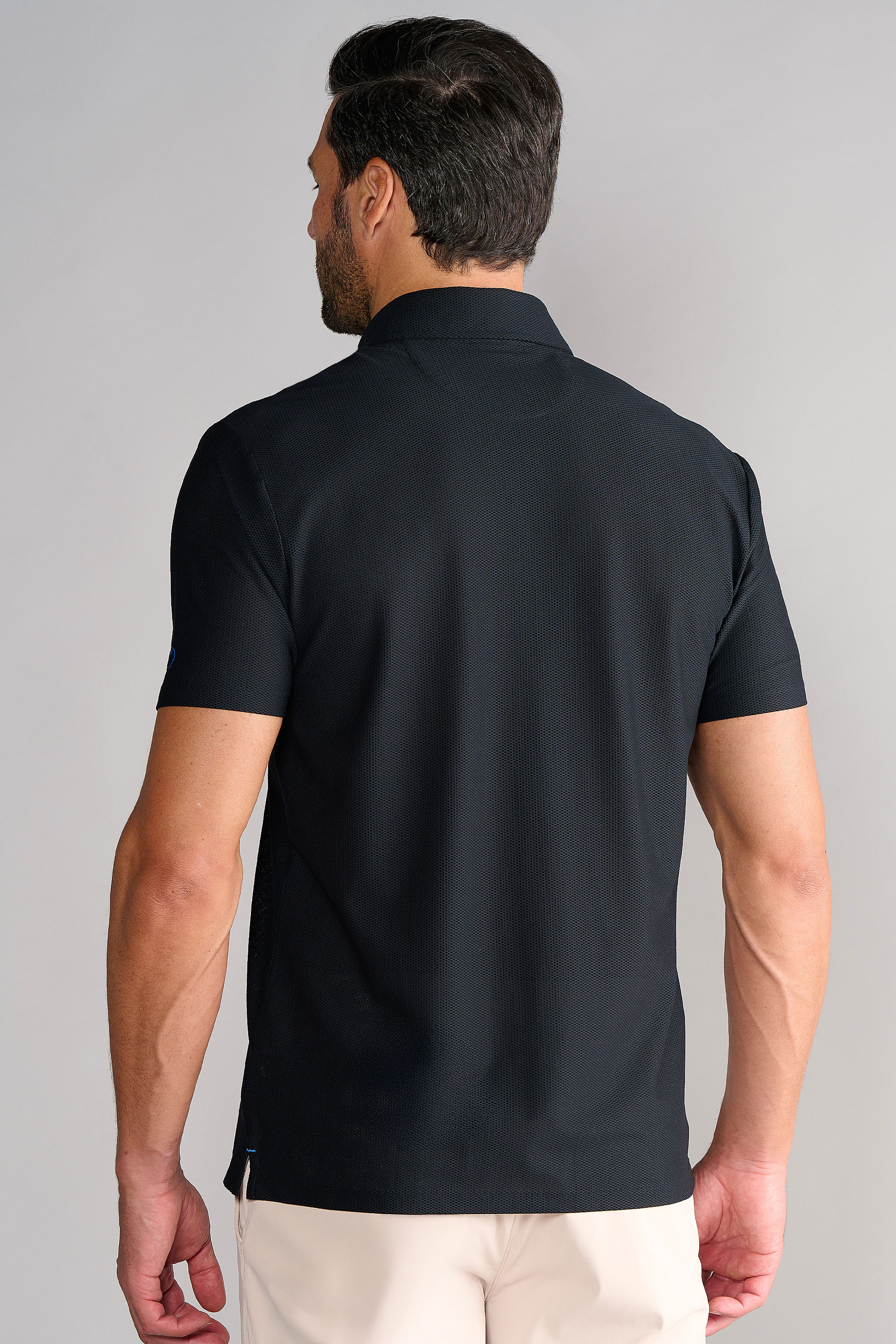 A man with short dark hair is seen from behind, wearing Twillory’s AIR 3 Button Polo Solid in Black and light beige pants. The upright collar and plain light gray background accentuate the polo’s tailored, modern design.