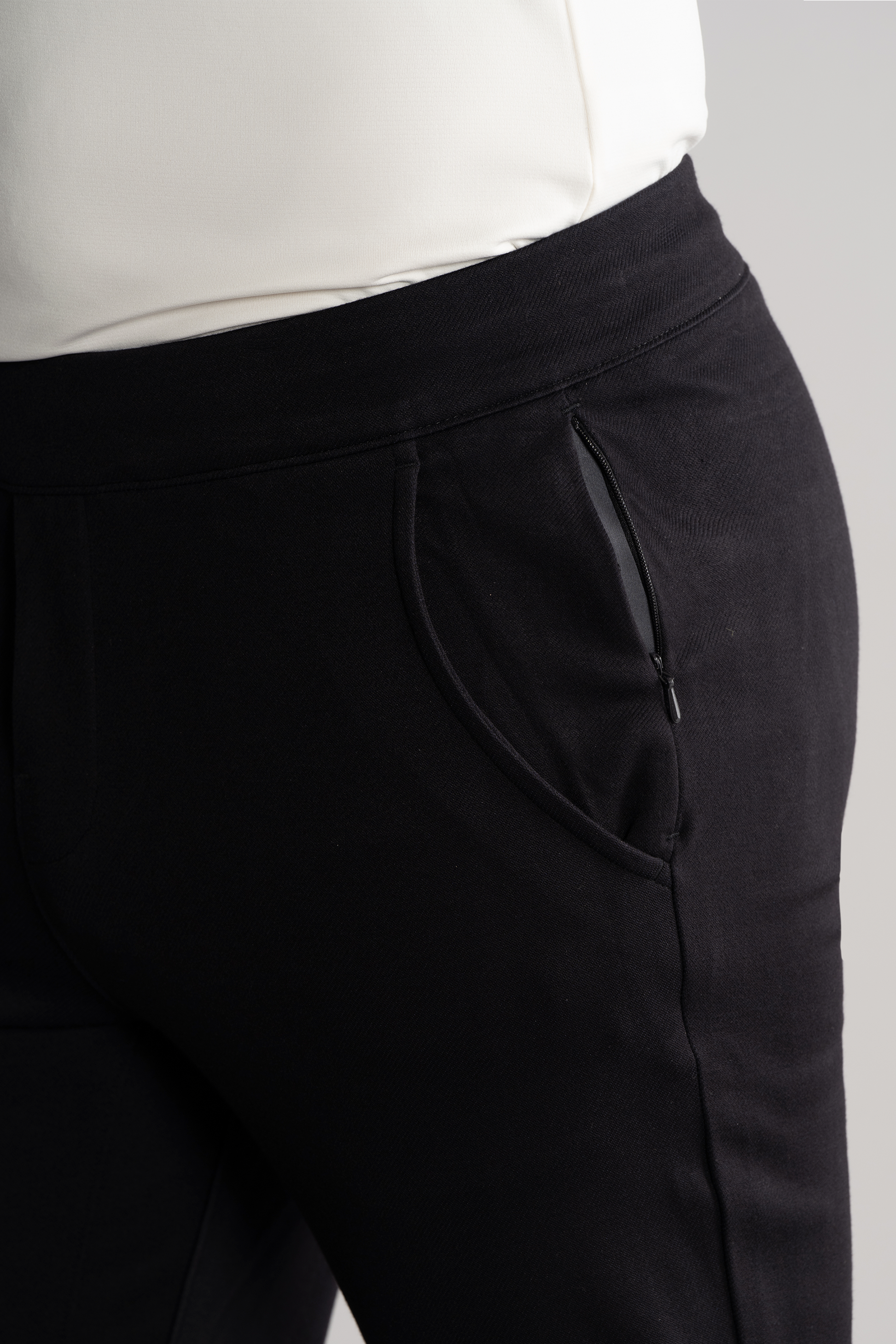 A close-up shows a person wearing Twillory’s Athlounger Jogger in black, featuring a side-zip pocket and comfort fabric. The focus is on the hip and upper thigh, paired with a white top against a neutral gray background.