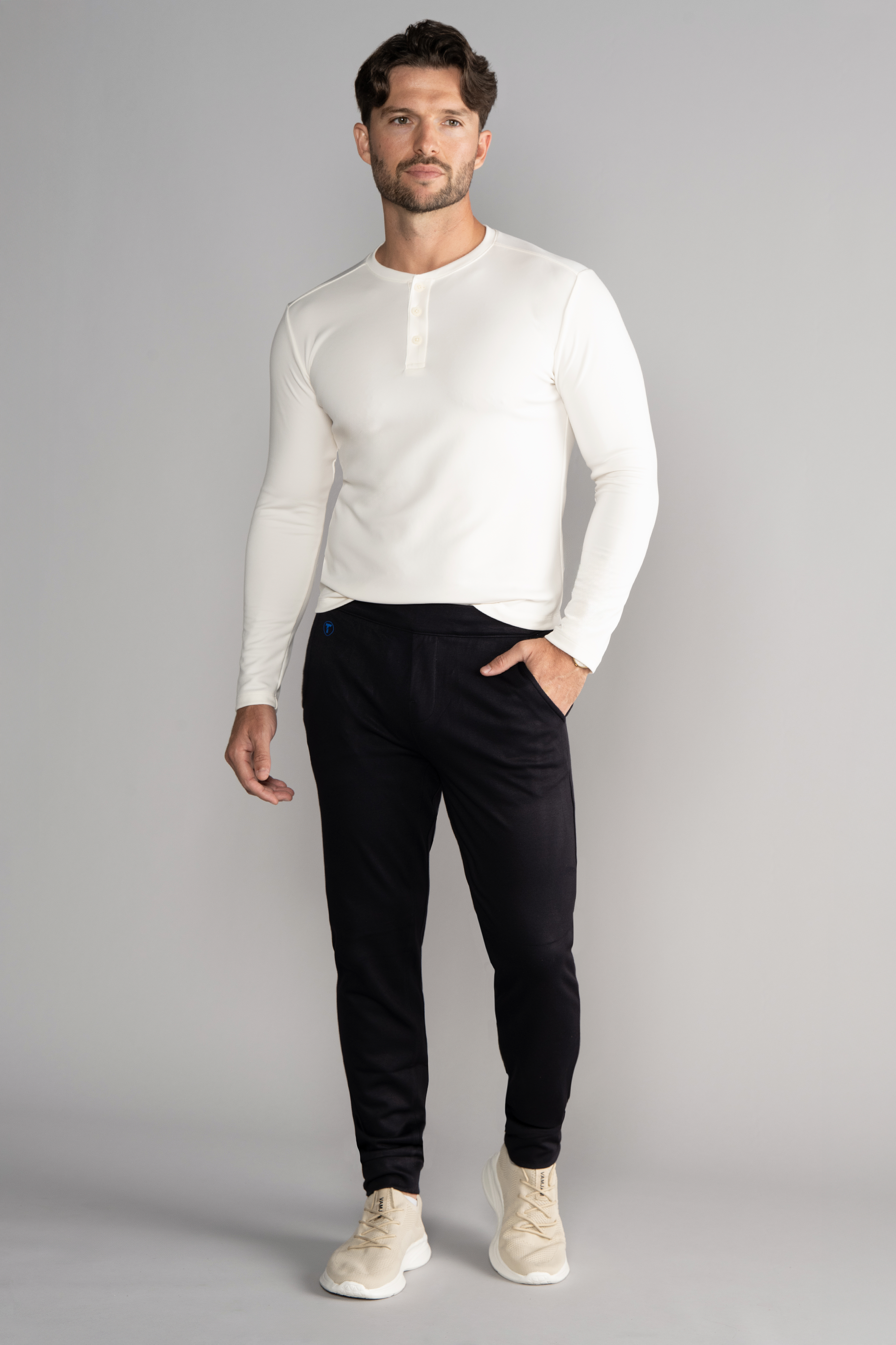 A man with short brown hair and a trimmed beard models the Twillory Athlounger Jogger, featuring a side-zip pocket, paired with a fitted white long-sleeve henley and beige sneakers. He faces forward with one hand in his pocket against a light gray background.