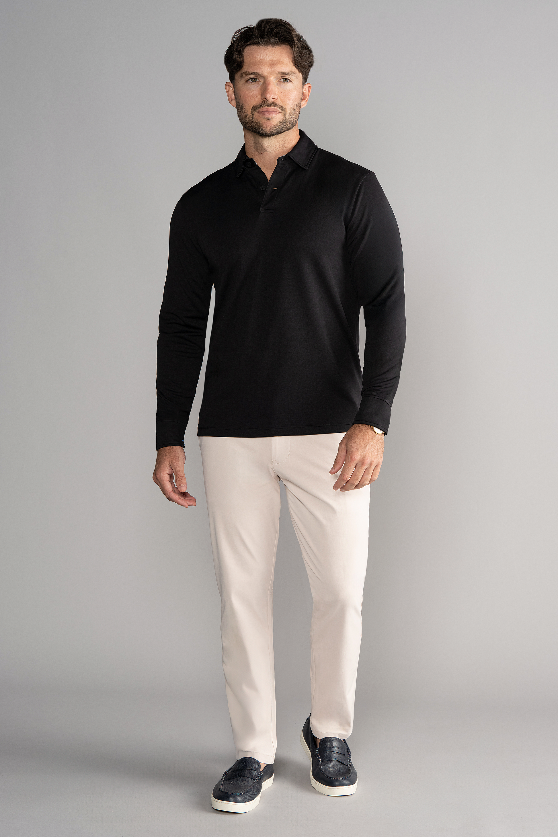 A man with short brown hair and a trimmed beard stands against a gray background wearing the Twillory Long Sleeve Polo Solid in black, light beige pants, and dark slip-on shoes. He has a neutral expression.