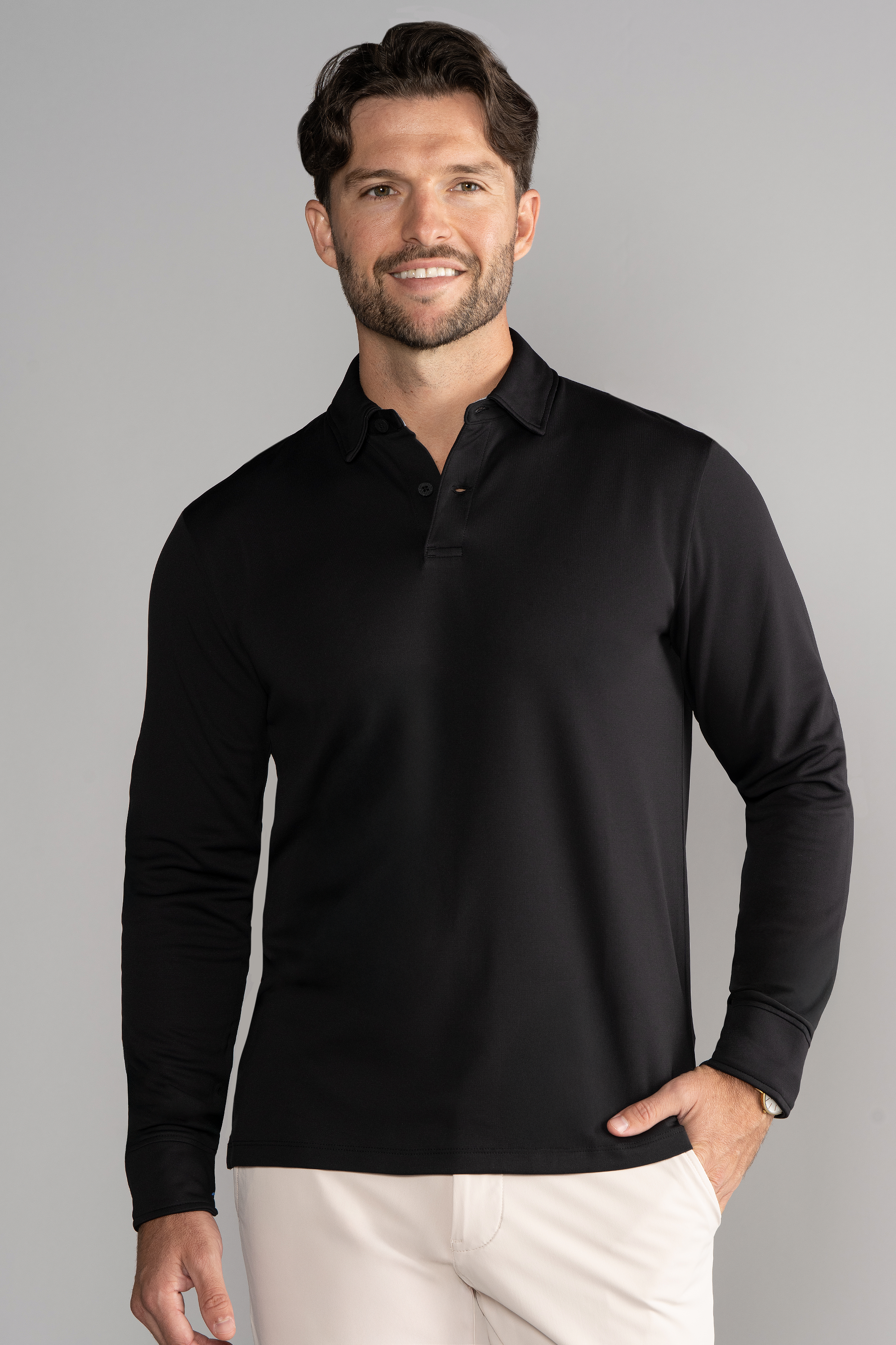 A man with short brown hair and a beard smiles at the camera, wearing the Twillory Long Sleeve Polo Solid in black and light beige pants. He stands against a plain gray background with one hand on his hip.