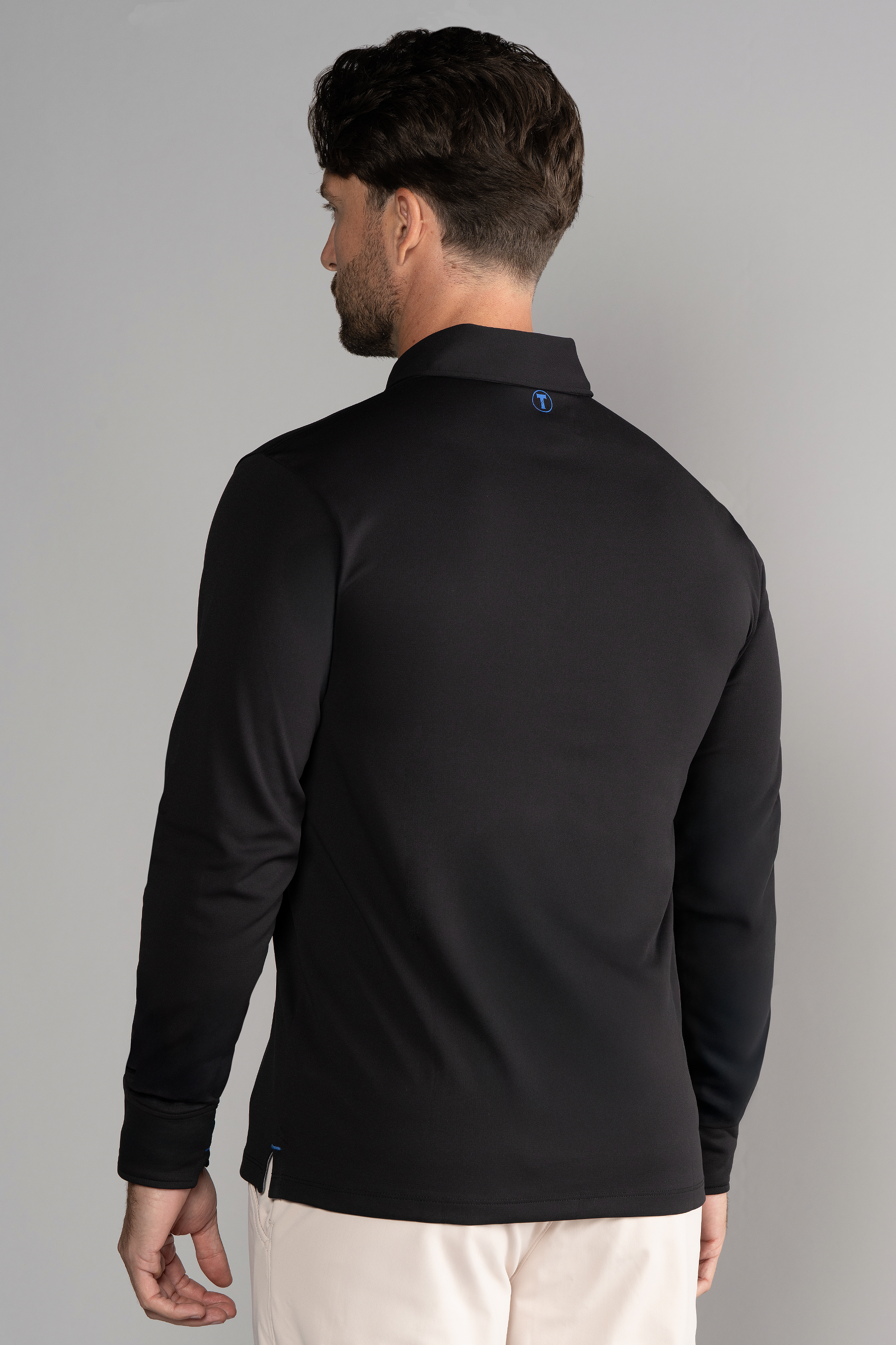 A man with short dark hair faces away from the camera, wearing the Twillory Long Sleeve Polo Solid in black. The plain gray background highlights a small blue logo at the upper back of his shirt and light-colored pants.
