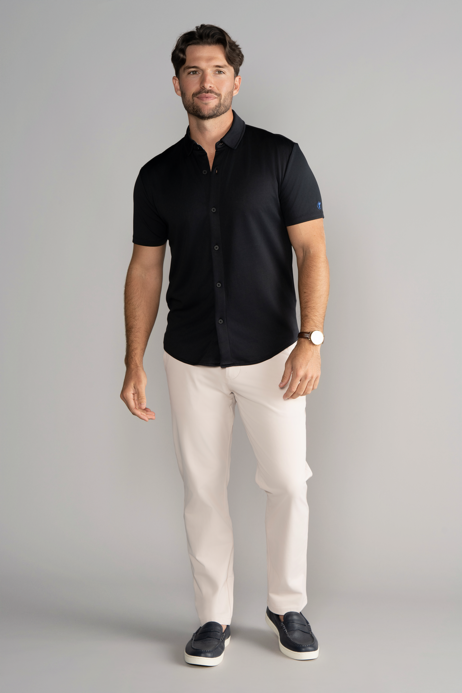 A man with short brown hair and a beard smiles against a gray background, wearing Twillory’s Short Sleeve Button Down Polo Solid in black, paired with white pants, dark slip-on shoes, and a watch. One arm is relaxed, the other slightly bent.