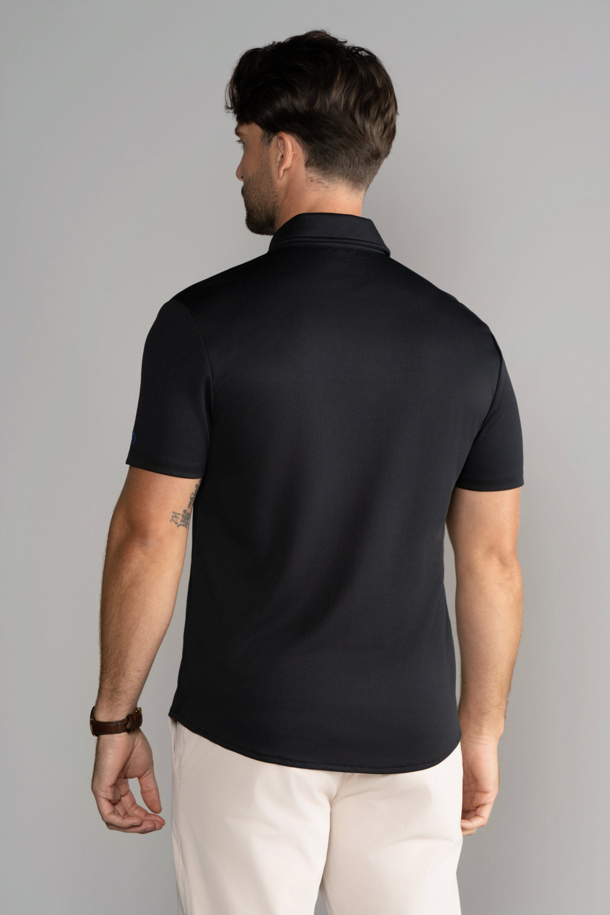 A man with short brown hair is seen from behind, wearing the Twillory Short Sleeve Button Down Polo Solid in Black and light beige pants. He has a brown watch on his left wrist, a small tattoo on his upper left arm, and stands against a light gray background.