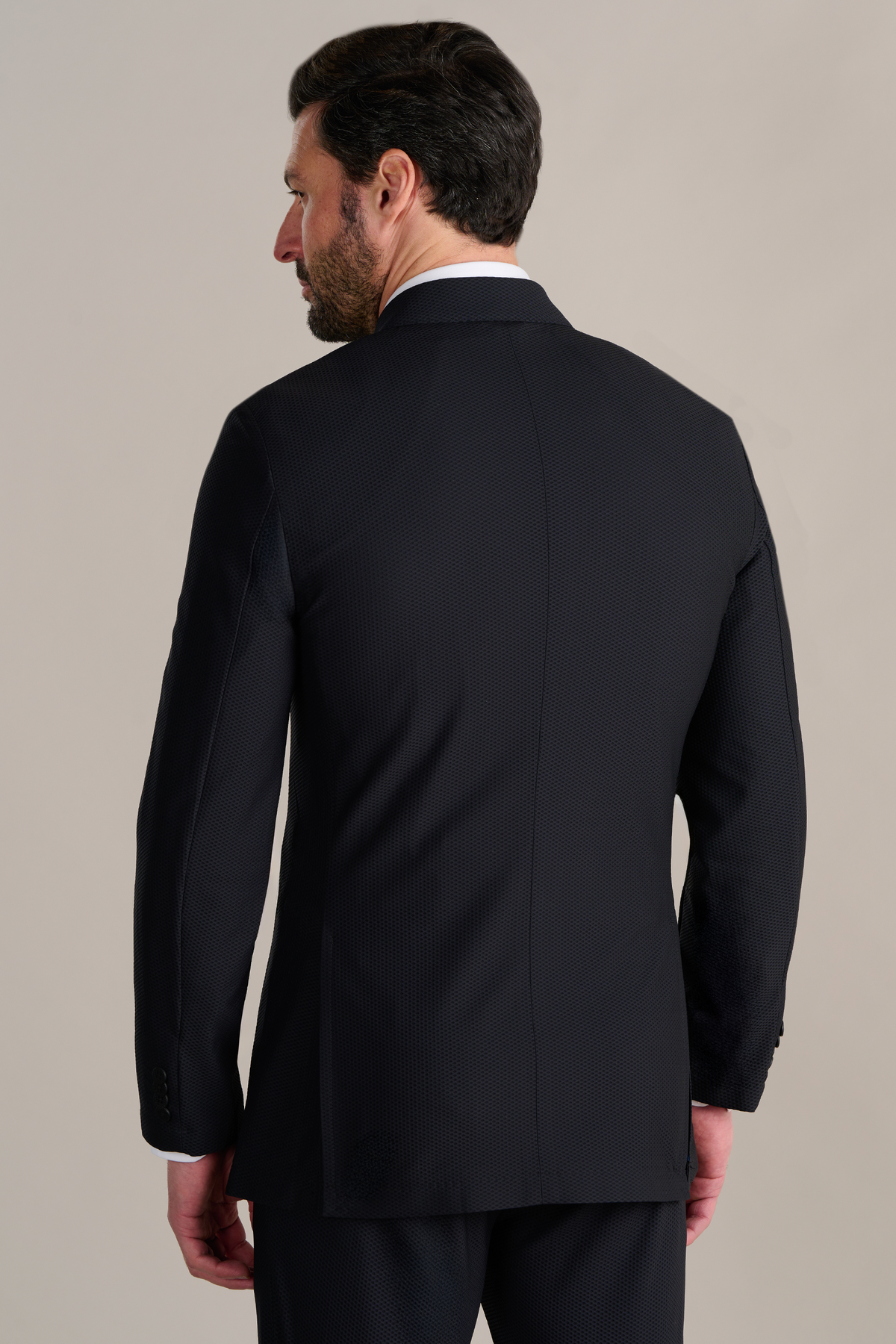 A man with dark hair and a beard is seen from behind wearing the Twillory AIR Double Breasted Blazer in Black over a white dress shirt, standing against a plain light gray background and looking to his left.