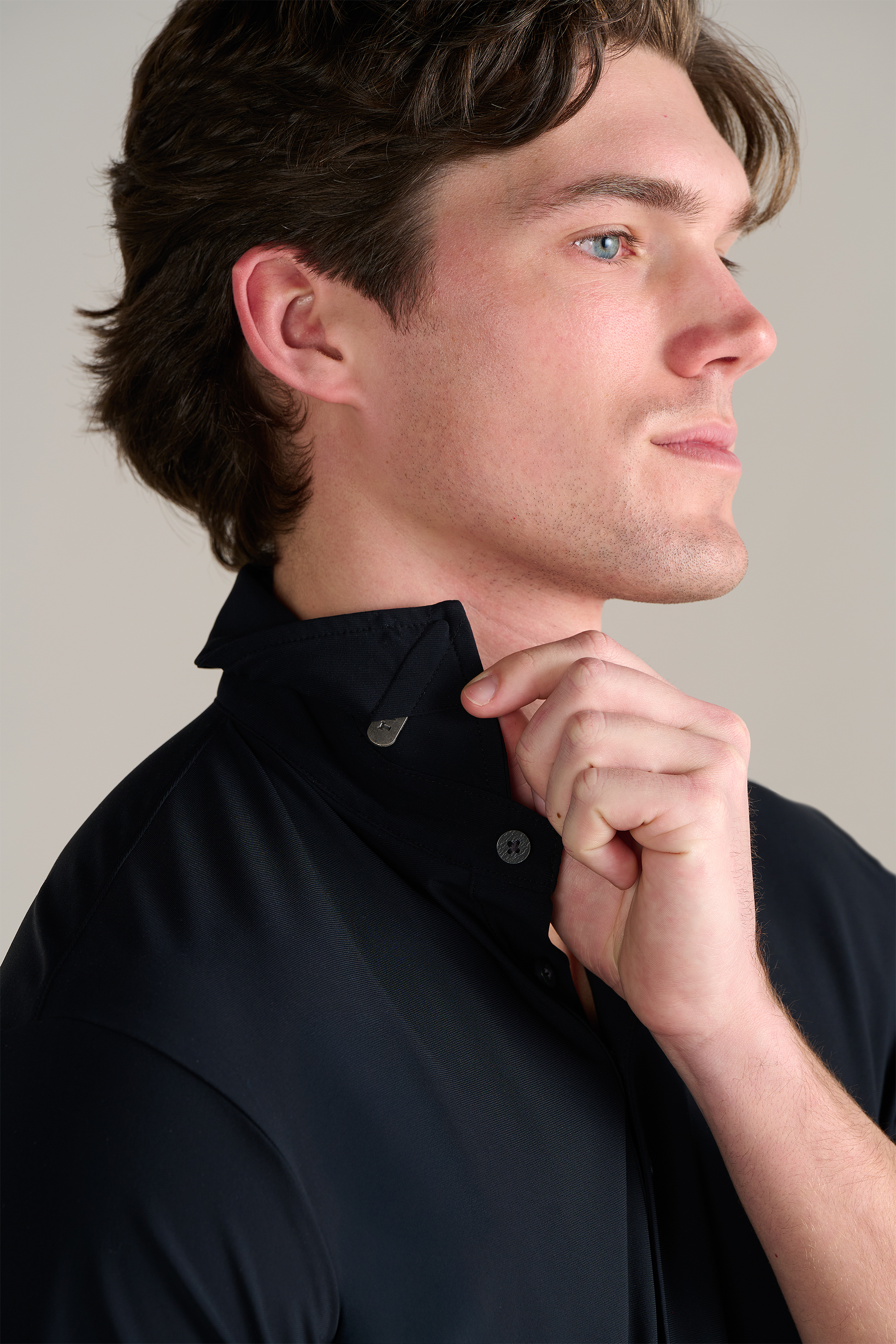 A light-skinned man with brown hair faces right, gently holding the collar of his Twillory Event Polo Solid in Black. The shirt features a wrinkle-free finish and small metal collar buttons. The background is neutral and uncluttered.