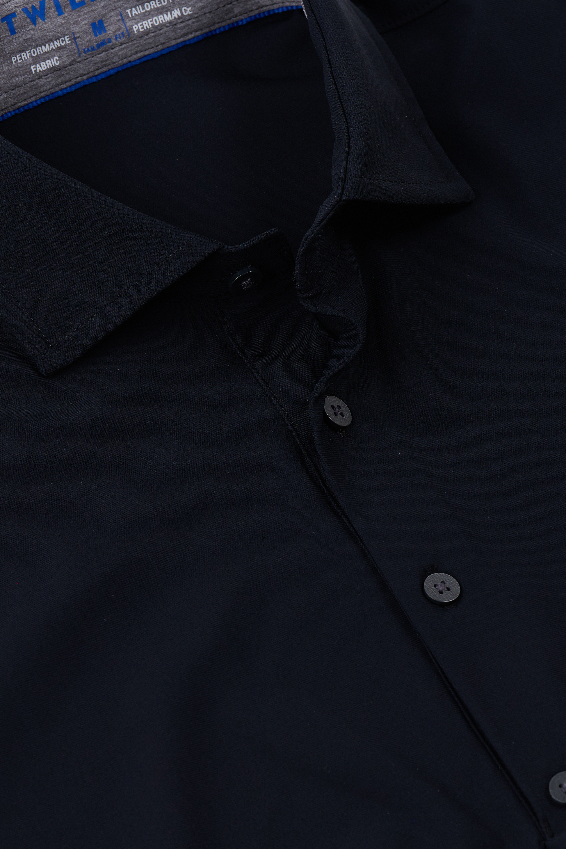Close-up of the Twillory Event Polo Solid in black, featuring a classic collar and black buttons. The performance fabric looks smooth and tailored, with blue text partially visible on the inner label inside the collar.