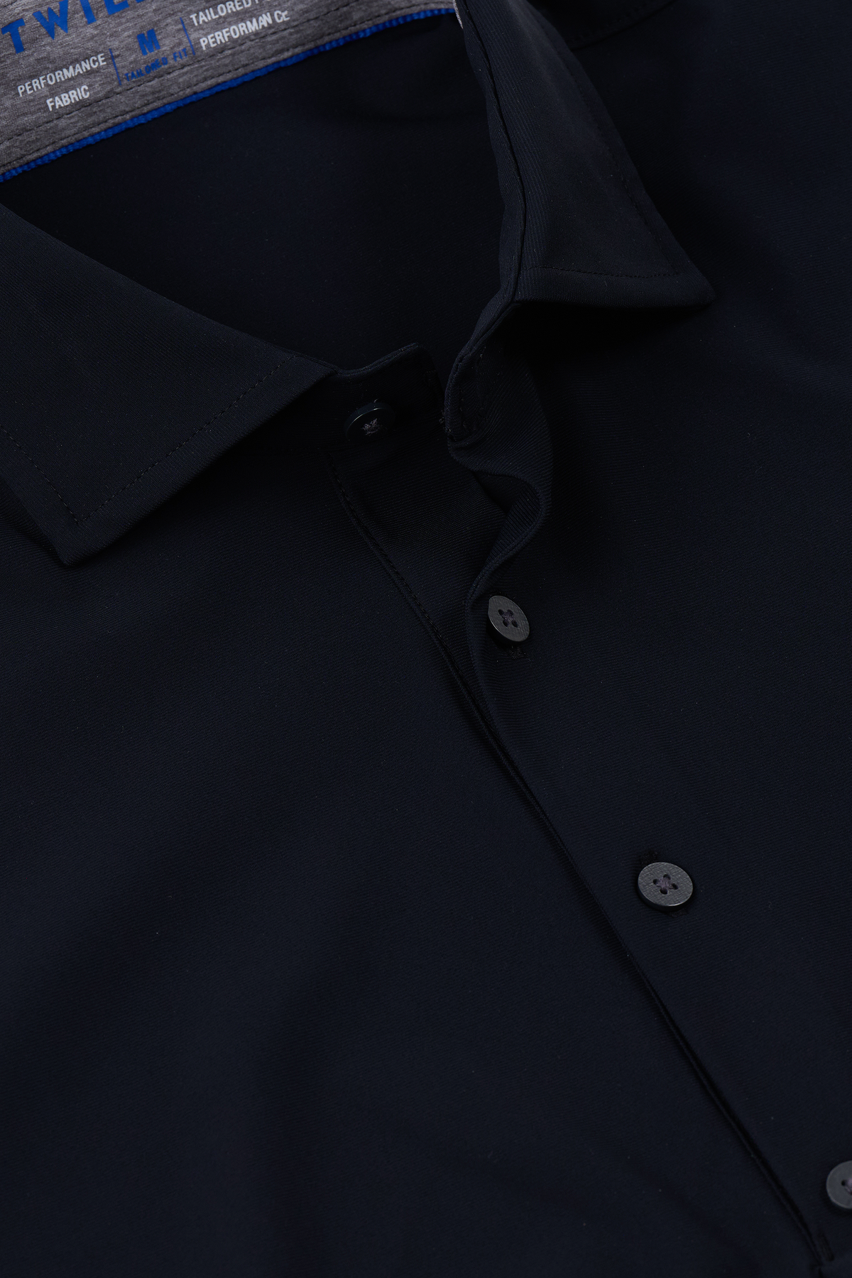 Close-up of the Twillory Event Polo Solid in black, featuring a classic collar and black buttons. The performance fabric looks smooth and tailored, with blue text partially visible on the inner label inside the collar.