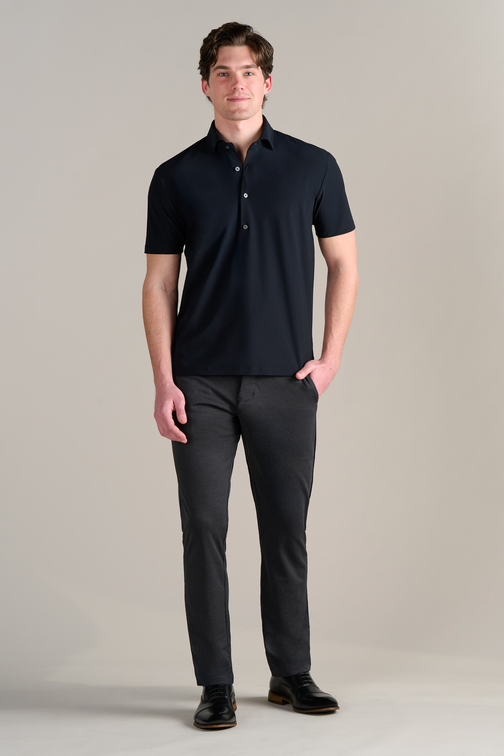 A man faces forward against a light-grey background, wearing the Twillory Event Polo Solid in black with a dress shirt collar, slim dark gray pants, and black dress shoes. He smiles slightly, one hand in his pocket and the other by his side.