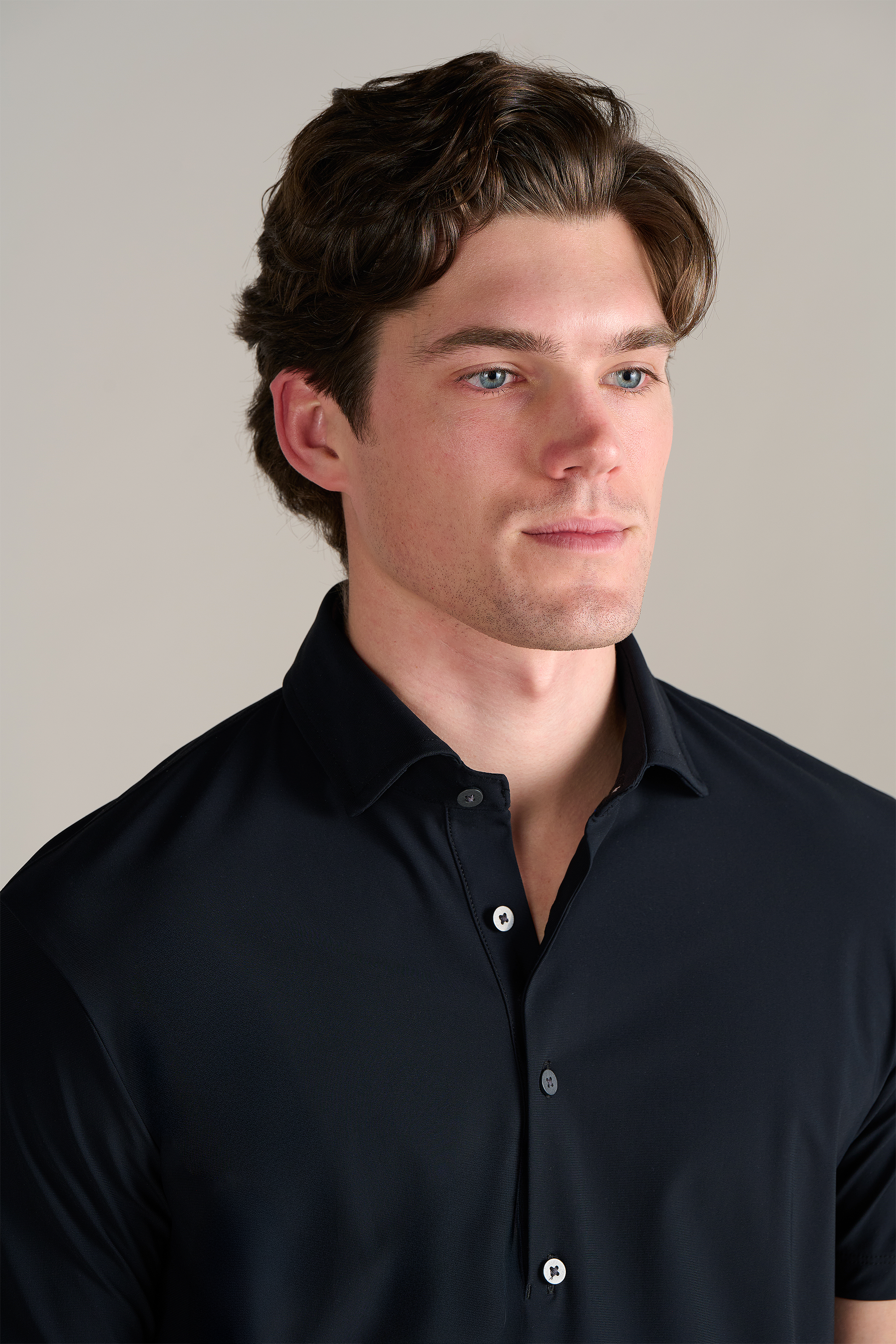 A young man with fair skin and short wavy brown hair, in a Twillory Event Polo Solid (Black), faces slightly to the side against a light background, drawing attention to his blue eyes and stylish attire.