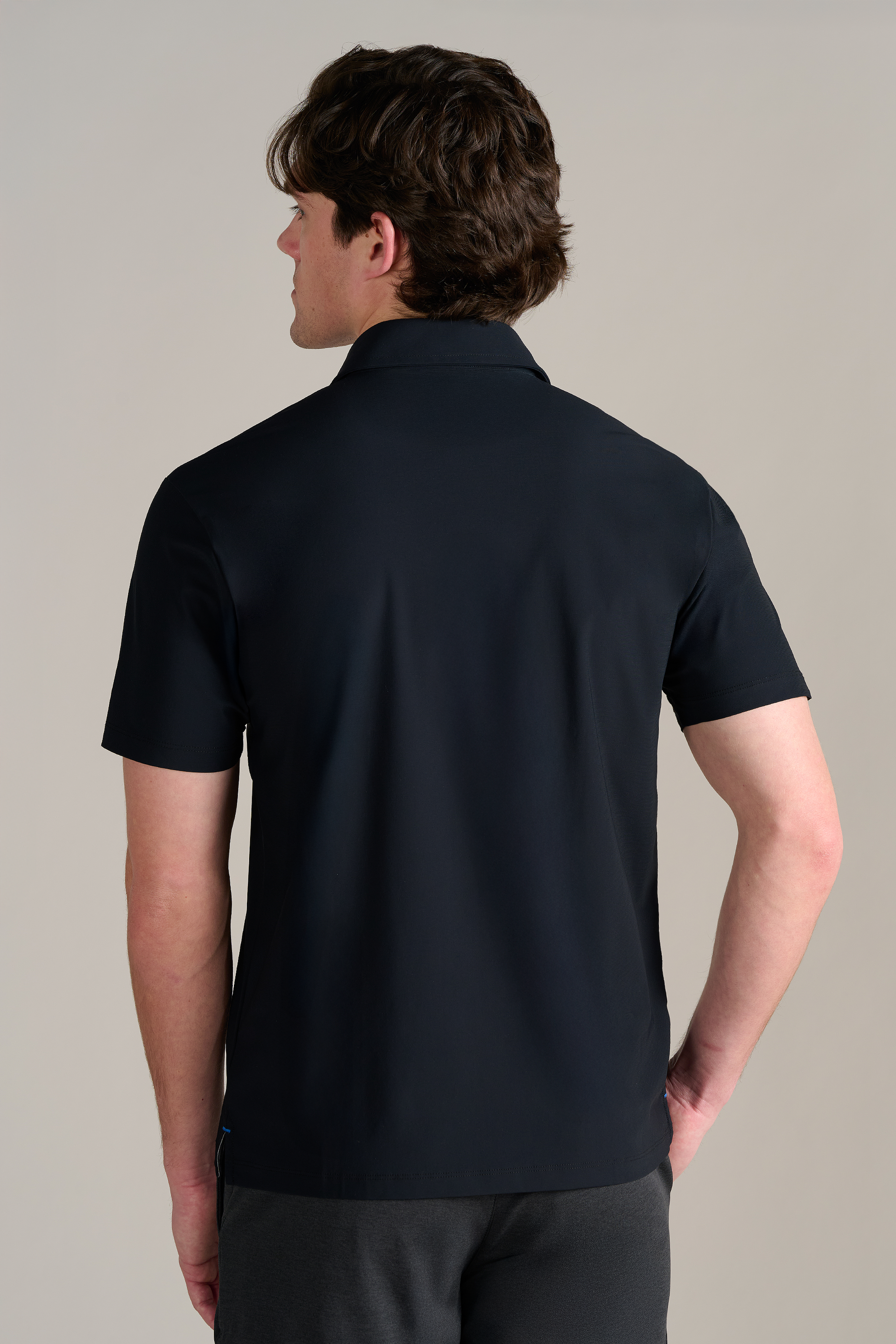 A man with short brown hair stands with his back to the camera, wearing Twillory’s Event Polo Solid in black and dark gray pants. The performance fabric polo features a dress shirt collar. His left hand is on his hip against a plain, light background.