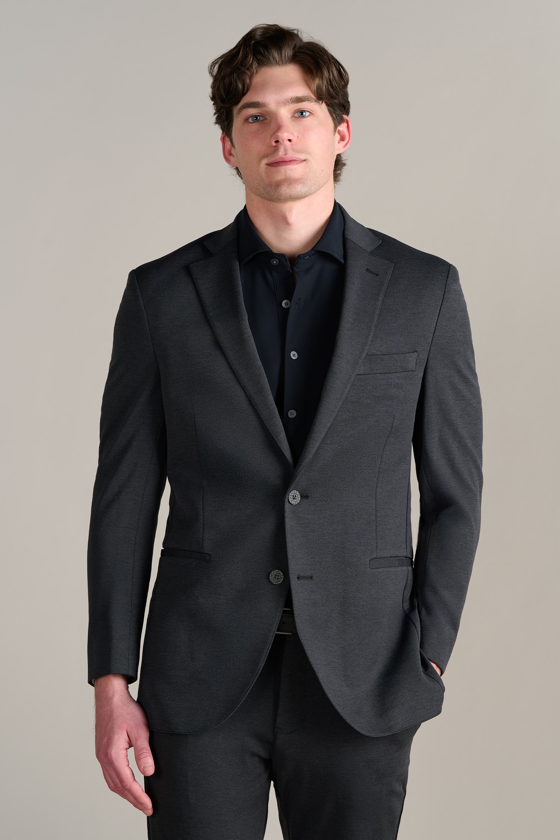 A young man with wavy brown hair wears Twillory’s Event Polo Solid in black, paired with a dark gray suit. He faces the camera against a plain light beige background, smiling slightly with his left hand in his pocket.