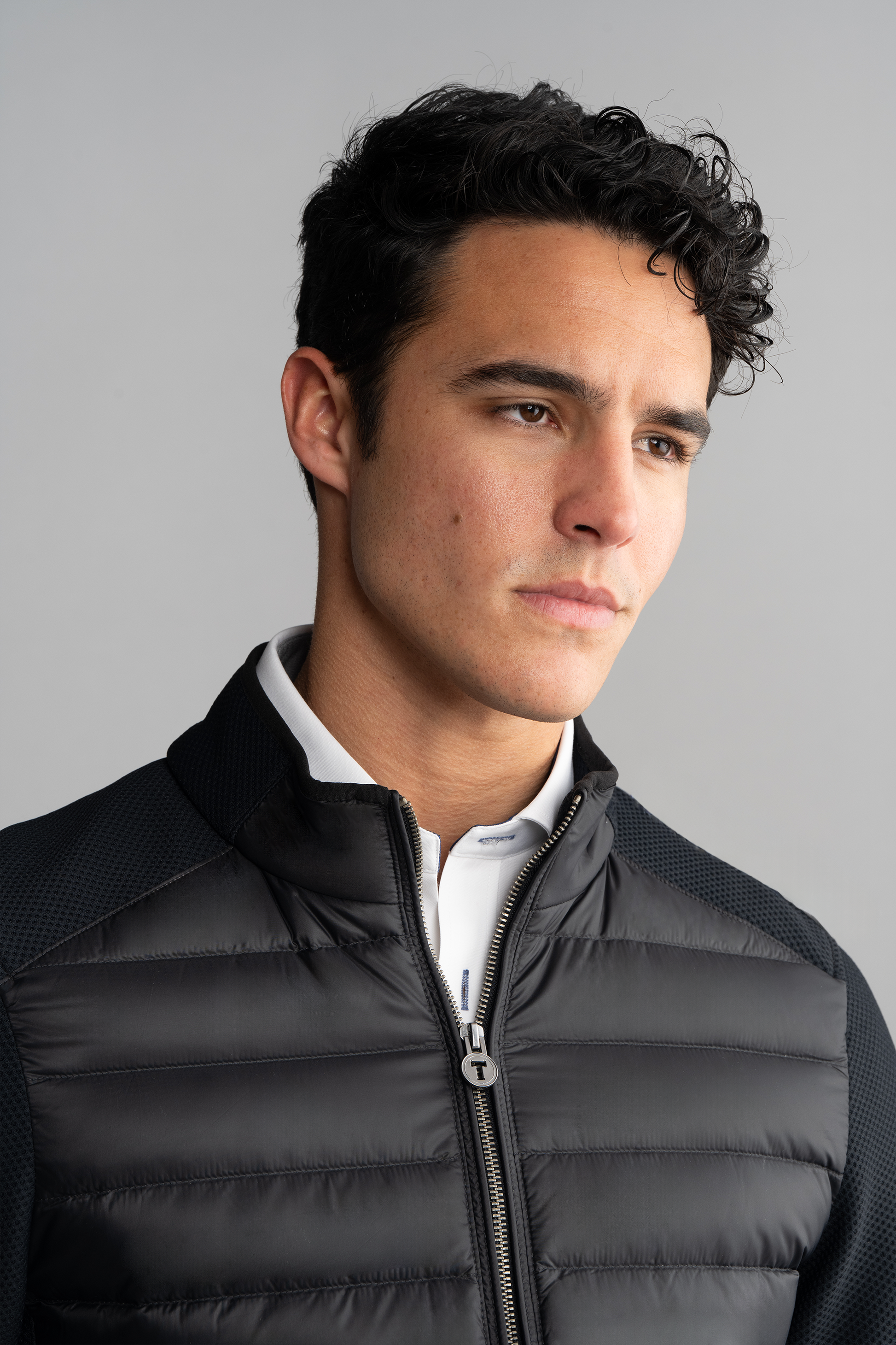 A young man with short, dark, curly hair and light skin wears the Twillory Central Hybrid Jacket in black over a white collared shirt. He looks slightly to the side with a neutral, pensive expression against a plain light gray background.