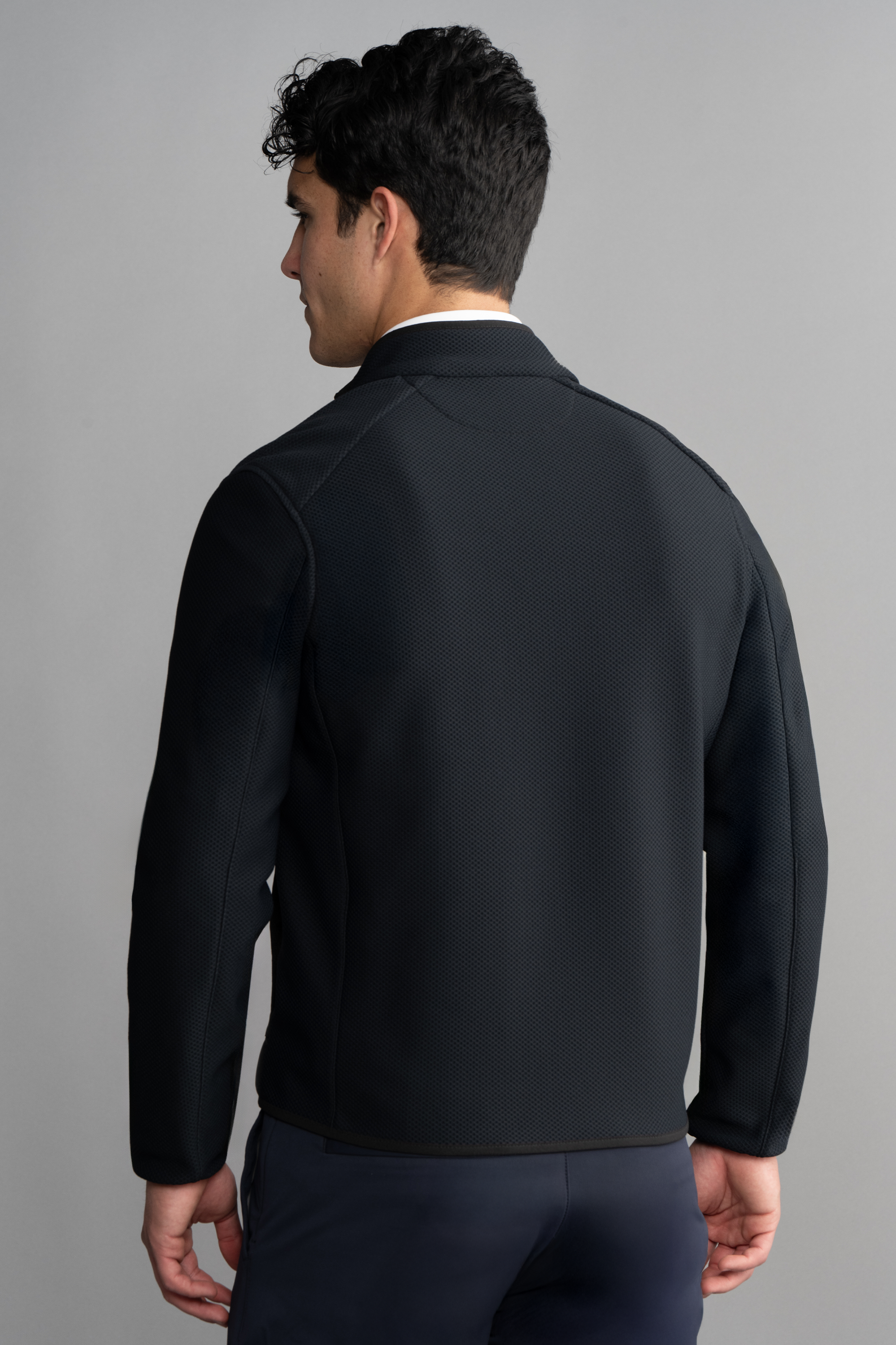 A man with short dark hair stands with his back to the camera, modeling the Twillory Central Hybrid Jacket in black against a plain gray background. His face is turned slightly to the side but remains unseen.