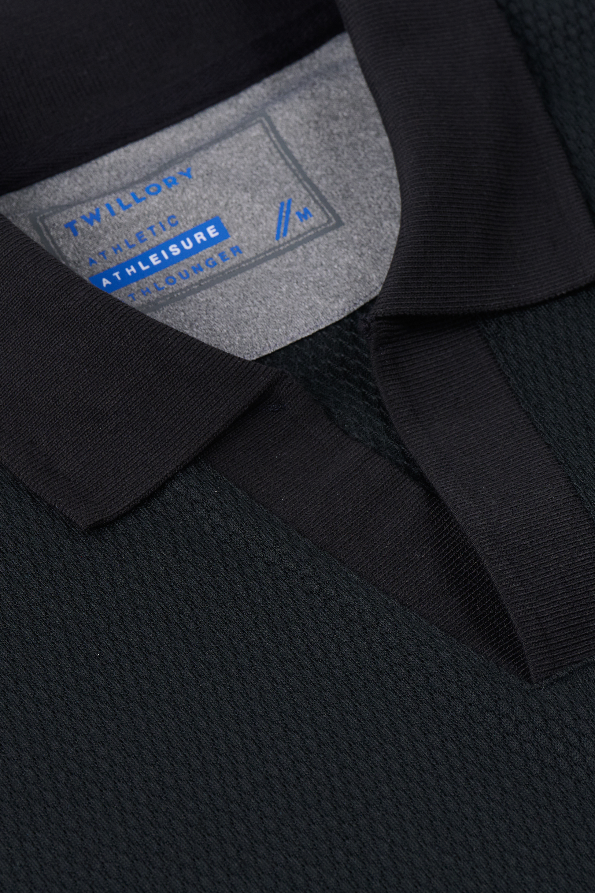 Close-up of the Twillory Long Sleeve Sweater Polo Solid in black, crafted from wrinkle-free performance knit. The inside label shows “Twillory Athletic Athleisure Concepts” in blue and grey, size M. Shirt is neatly folded to showcase collar and chest.