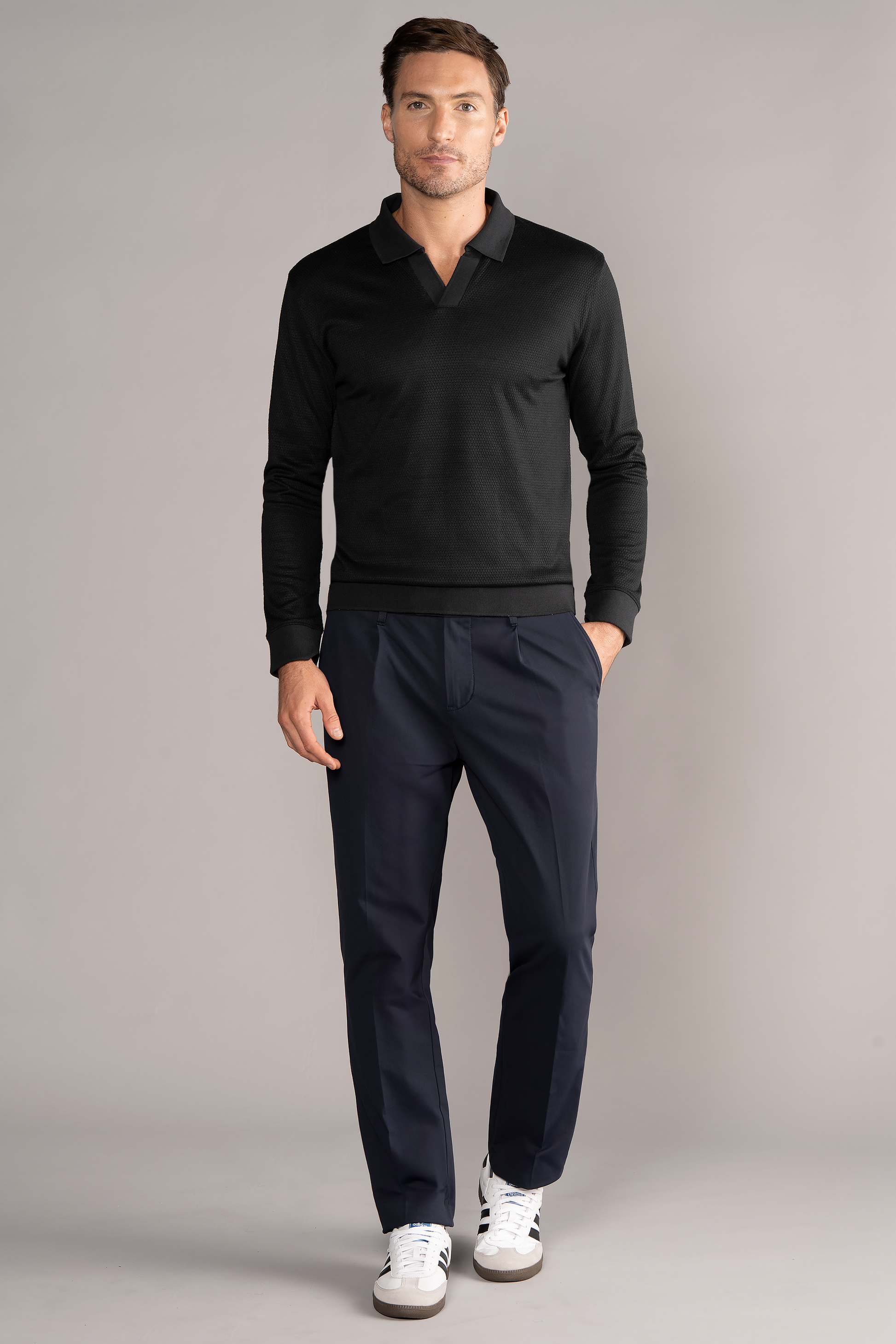 A man stands against a light gray background wearing the Twillory Long Sleeve Sweater Polo Solid in black, paired with navy tailored pants and white sneakers. One hand is in his pocket as he looks at the camera with a neutral expression.