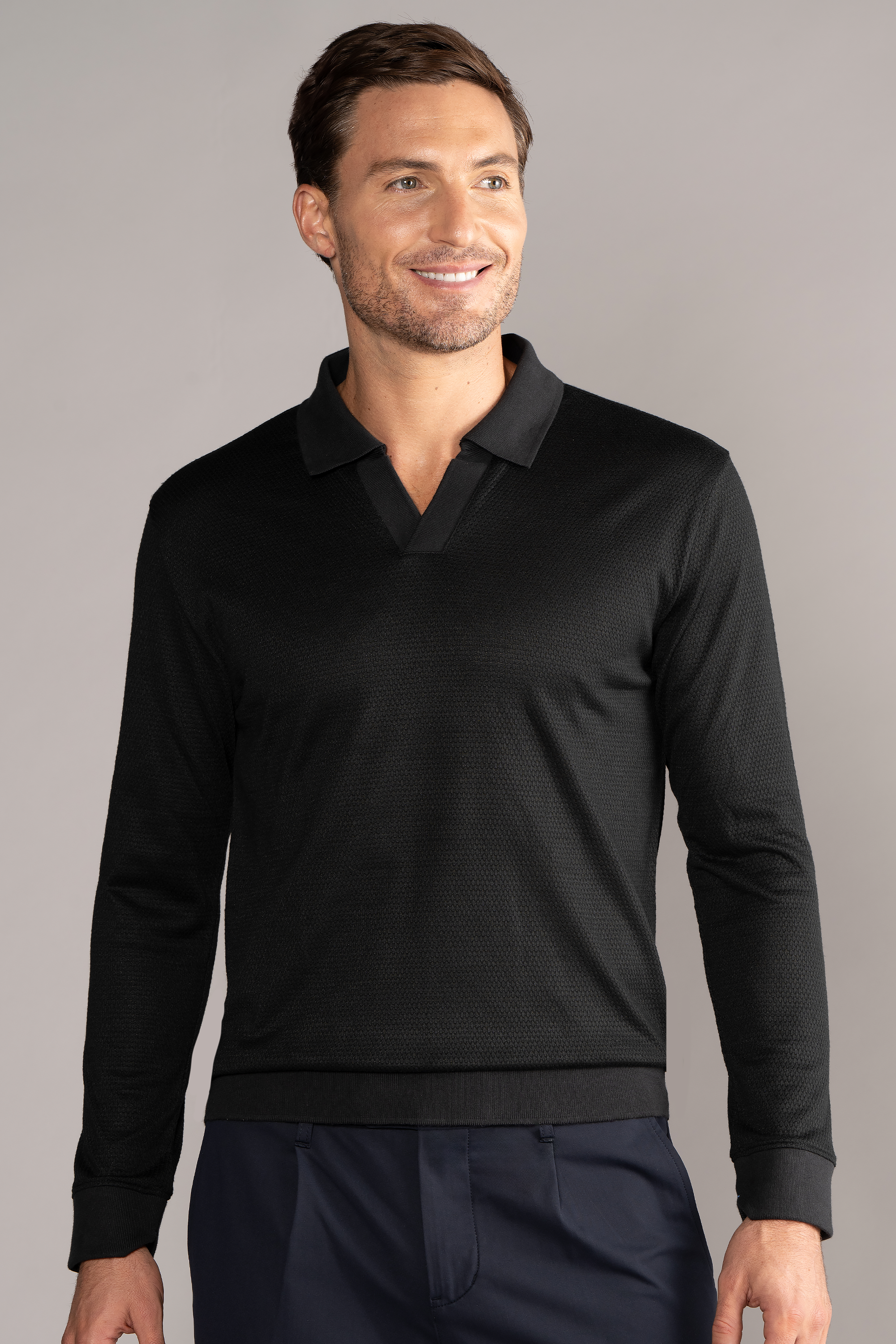 A man with short brown hair smiles while wearing the Twillory Long Sleeve Sweater Polo Solid in black, paired with dark navy pants, standing against a plain light gray background.