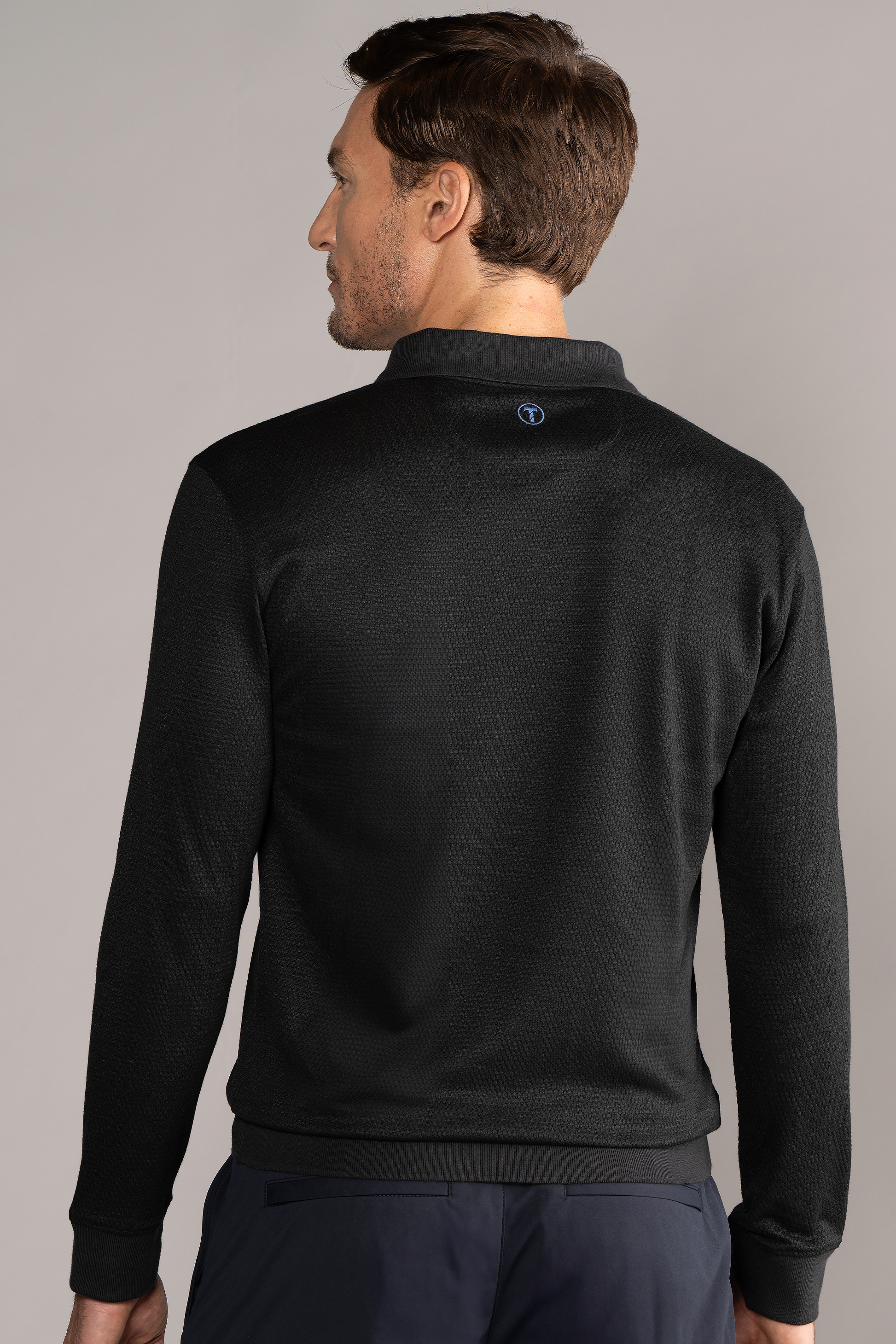 A man with short brown hair faces away, wearing the Twillory Long Sleeve Sweater Polo Solid in black, featuring a small circular blue and white logo below the collar. He stands against a plain gray background and also wears navy pants.