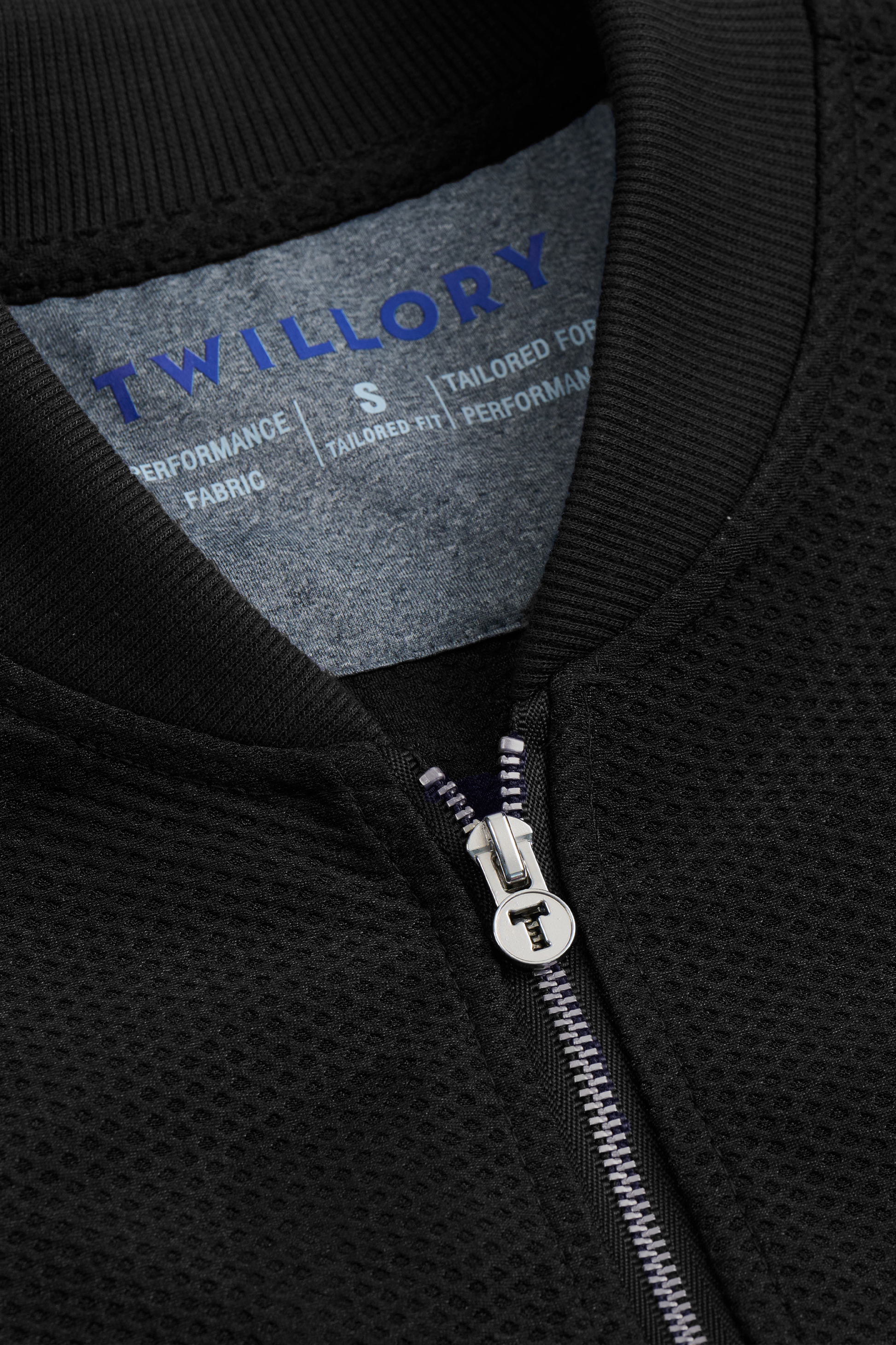 Close-up of the Twillory AIR Bomber // Black: this stretch bomber jacket has a textured, breathable outer, a silver zipper with branded pull, and an inside tag reading "Twillory" in blue with sizing and fabric details in white on grey.