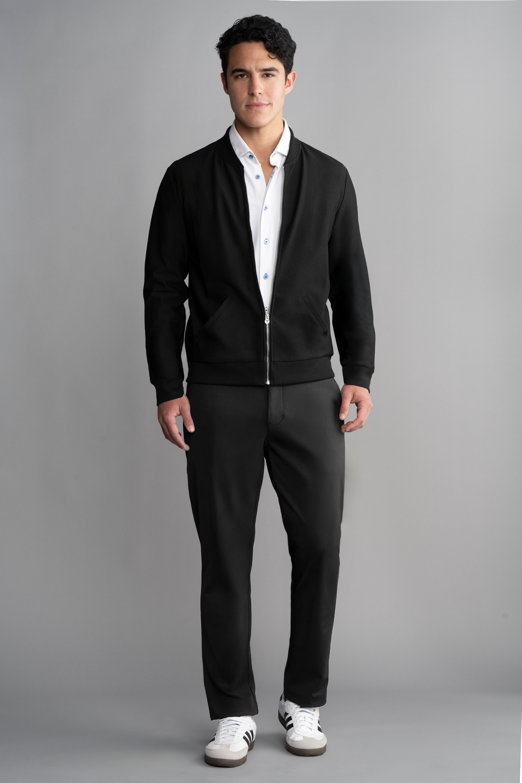 A man with short dark hair and a neutral expression stands against a gray background wearing Twillory's AIR Bomber in black over a white shirt, black pants, and white Adidas sneakers with black stripes. His arms are relaxed at his sides.