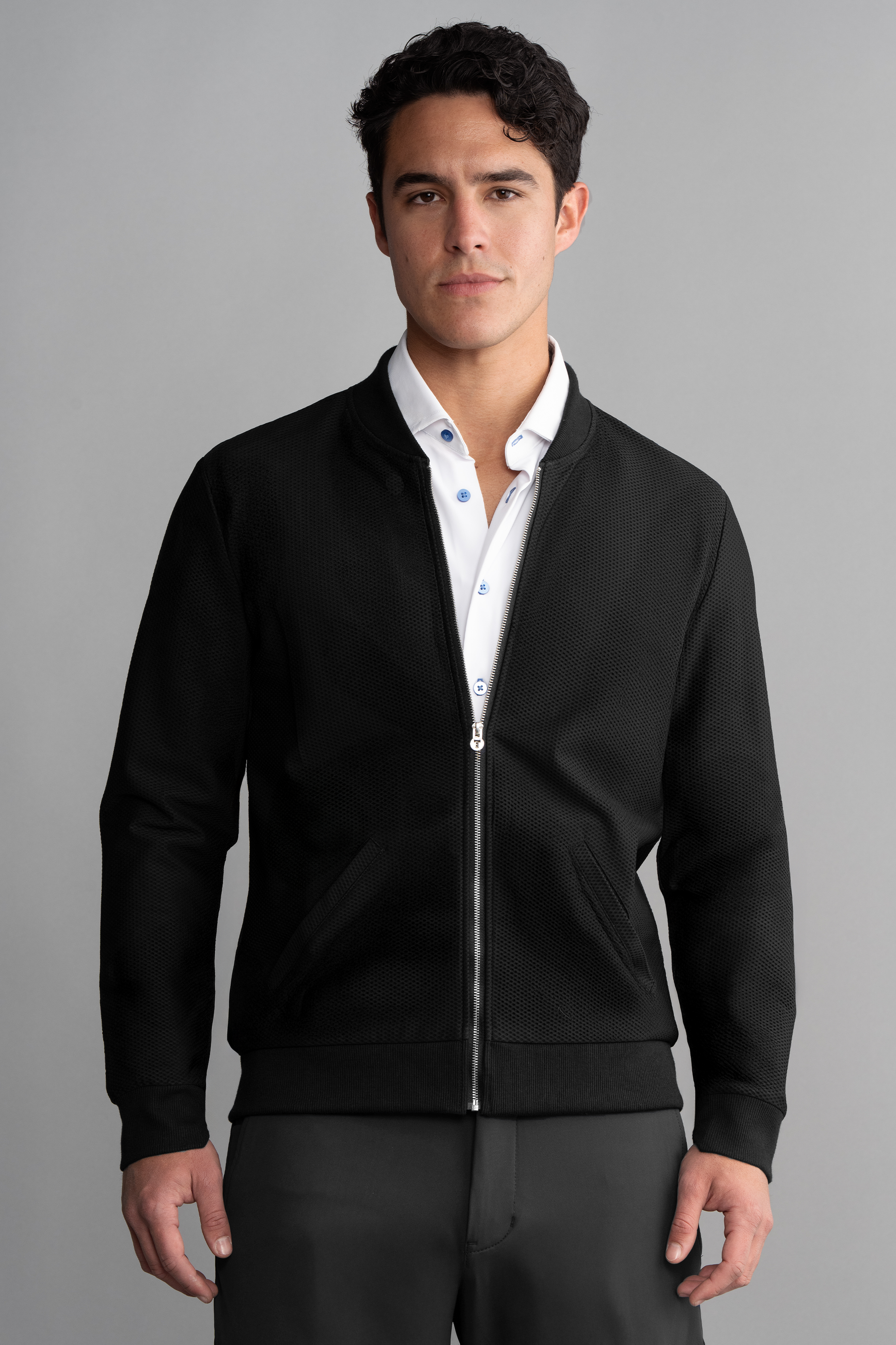 A man with short dark hair and a neutral expression wears the Twillory AIR Bomber in black over a white collared shirt with blue buttons, standing against a plain gray background.